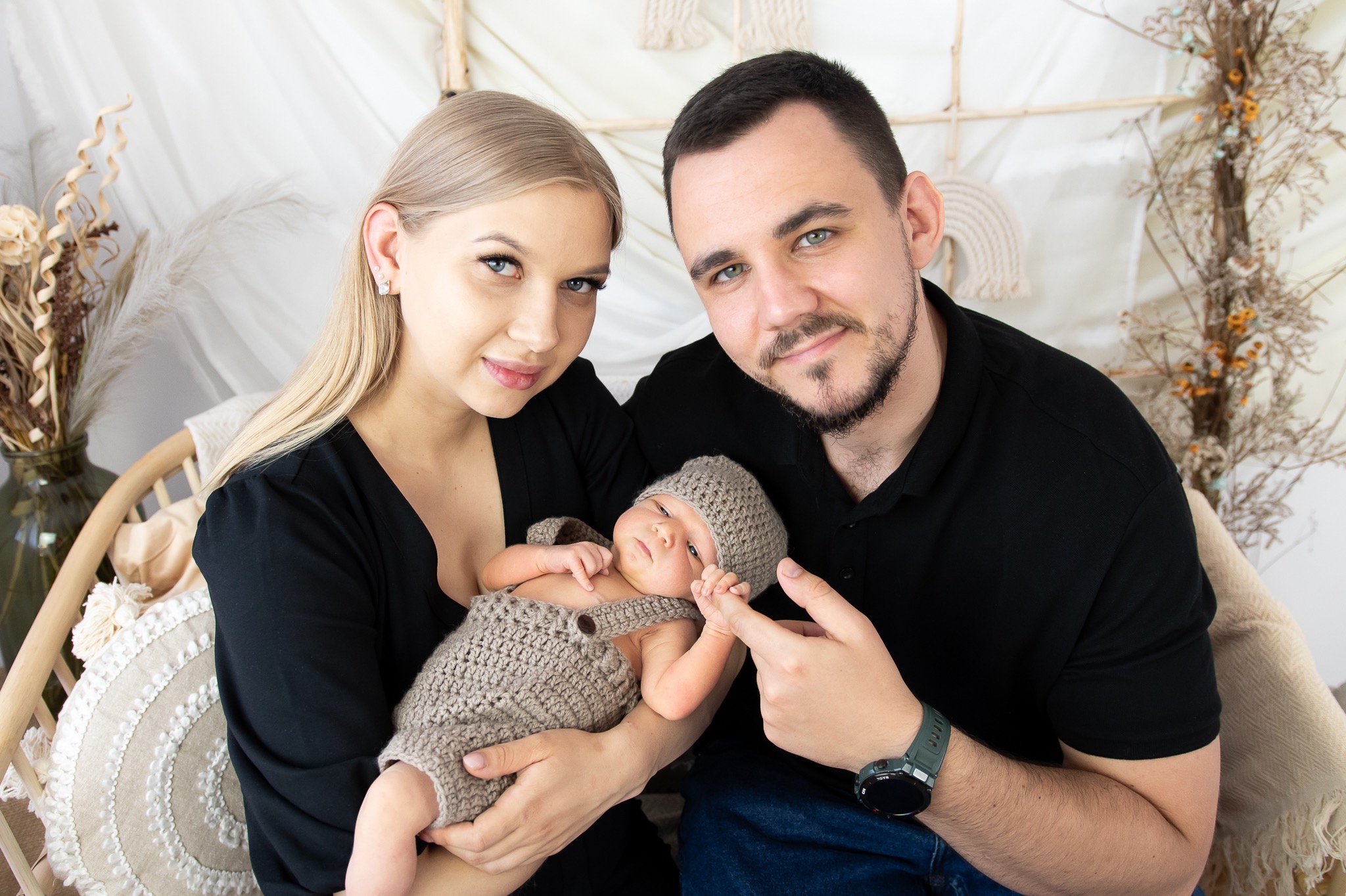 Rodzinne ujęcie: młoda para trzyma noworodka ubranego w dziergany kombinezon i czapeczkę, ojciec delikatnie dotyka paluszka dziecka, tło stanowią dekoracje w stylu boho.