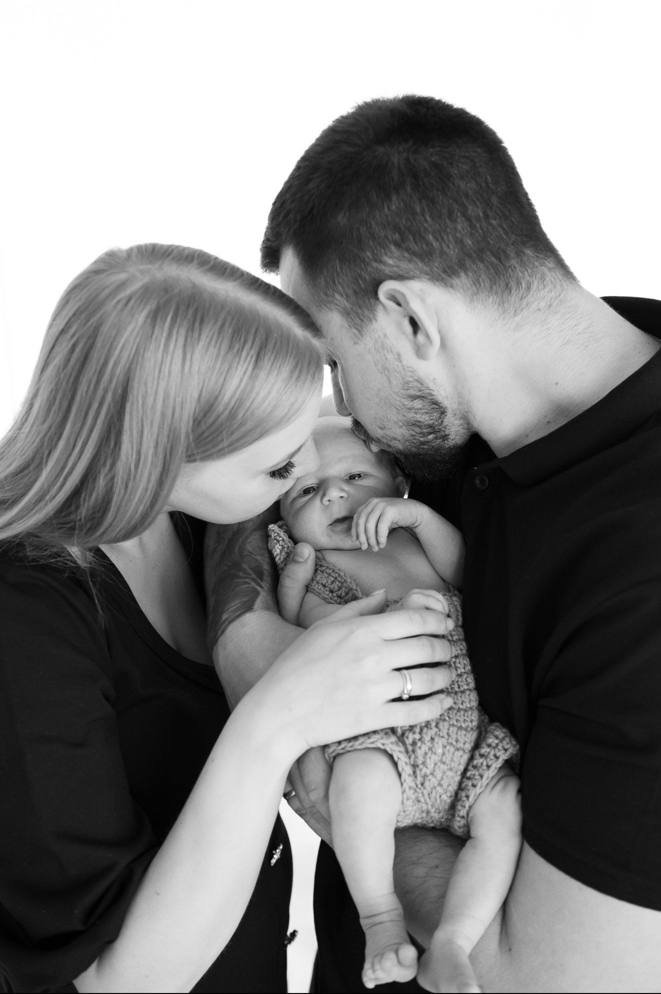 Czarno-białe zdjęcie rodziców całujących noworodka ubranego w dziergany kombinezon, z delikatnym uśmiechem na twarzy dziecka.