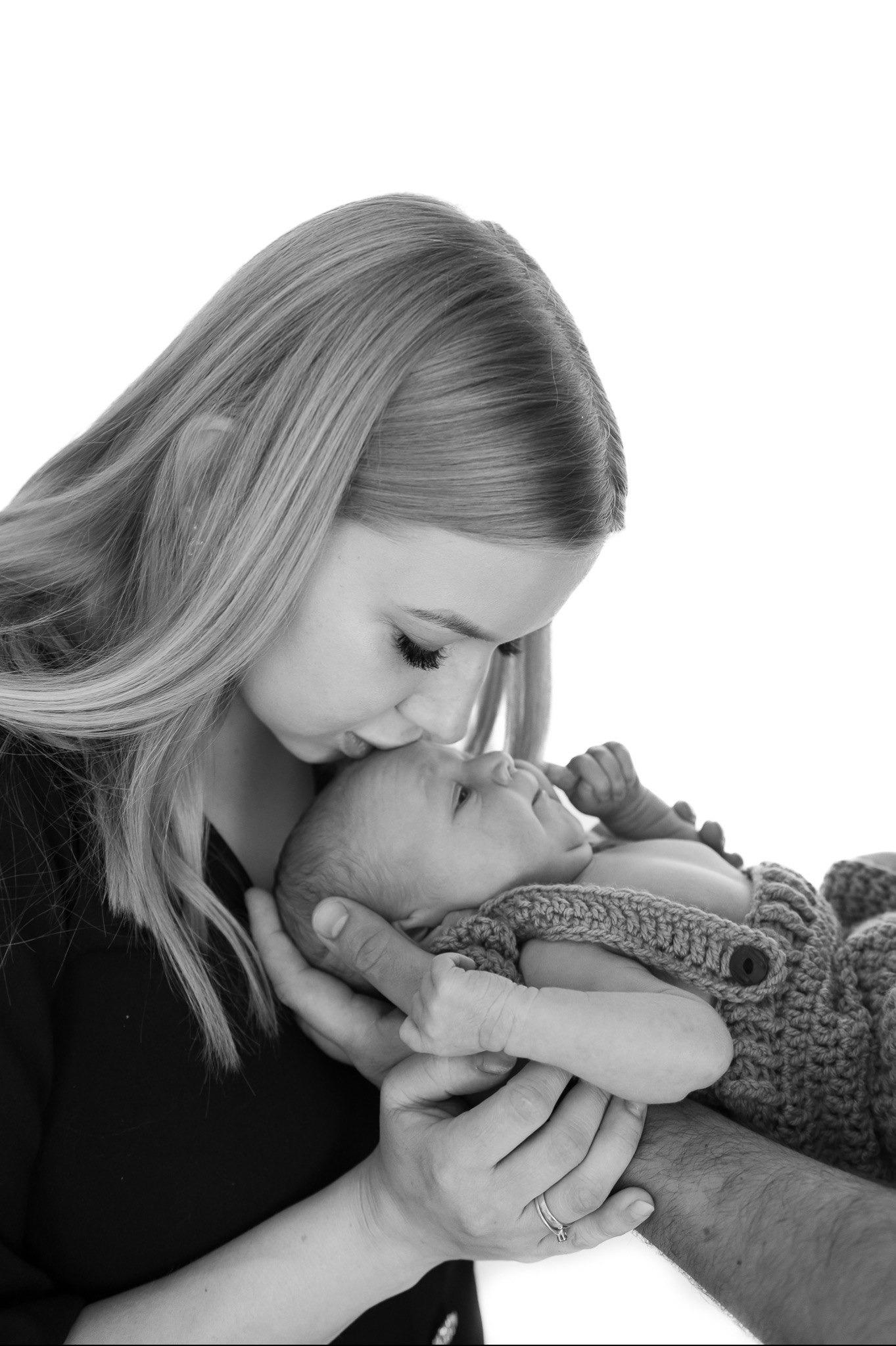 Czarno-białe zdjęcie matki całującej w czoło nowo narodzone dziecko, ubrane w dziergany kombinezon z guzikiem, trzymane w dłoniach obojga rodziców.
