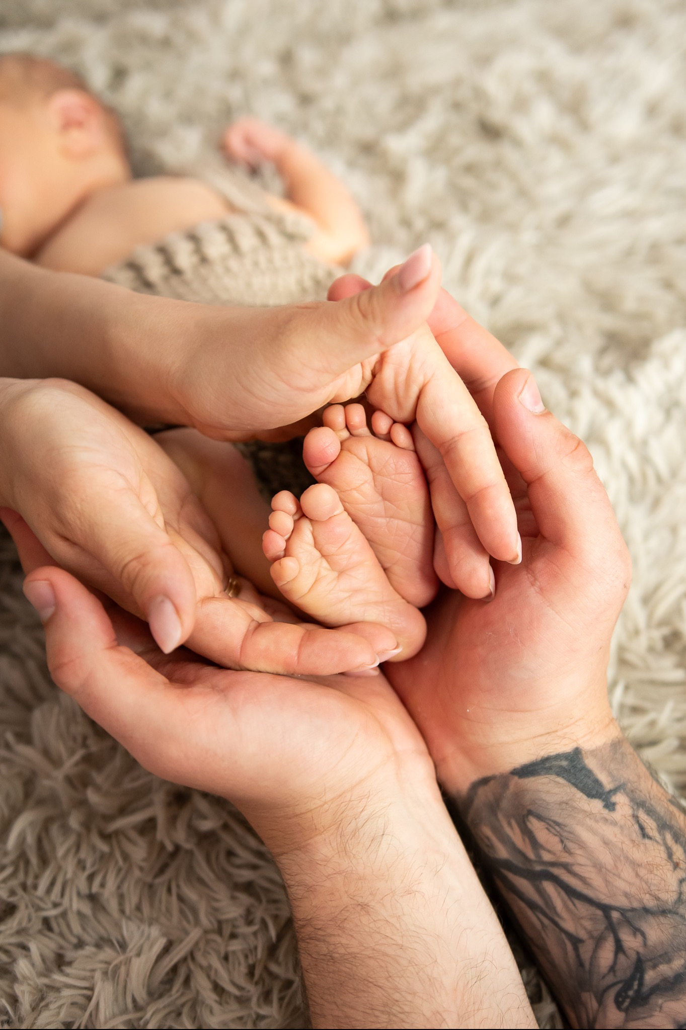 Dłonie rodziców trzymające stopy noworodka na puszystym, beżowym tle, widoczny fragment ciała dziecka owiniętego w dzianinowy kocyk, tatuaż na przedramieniu ojca.
