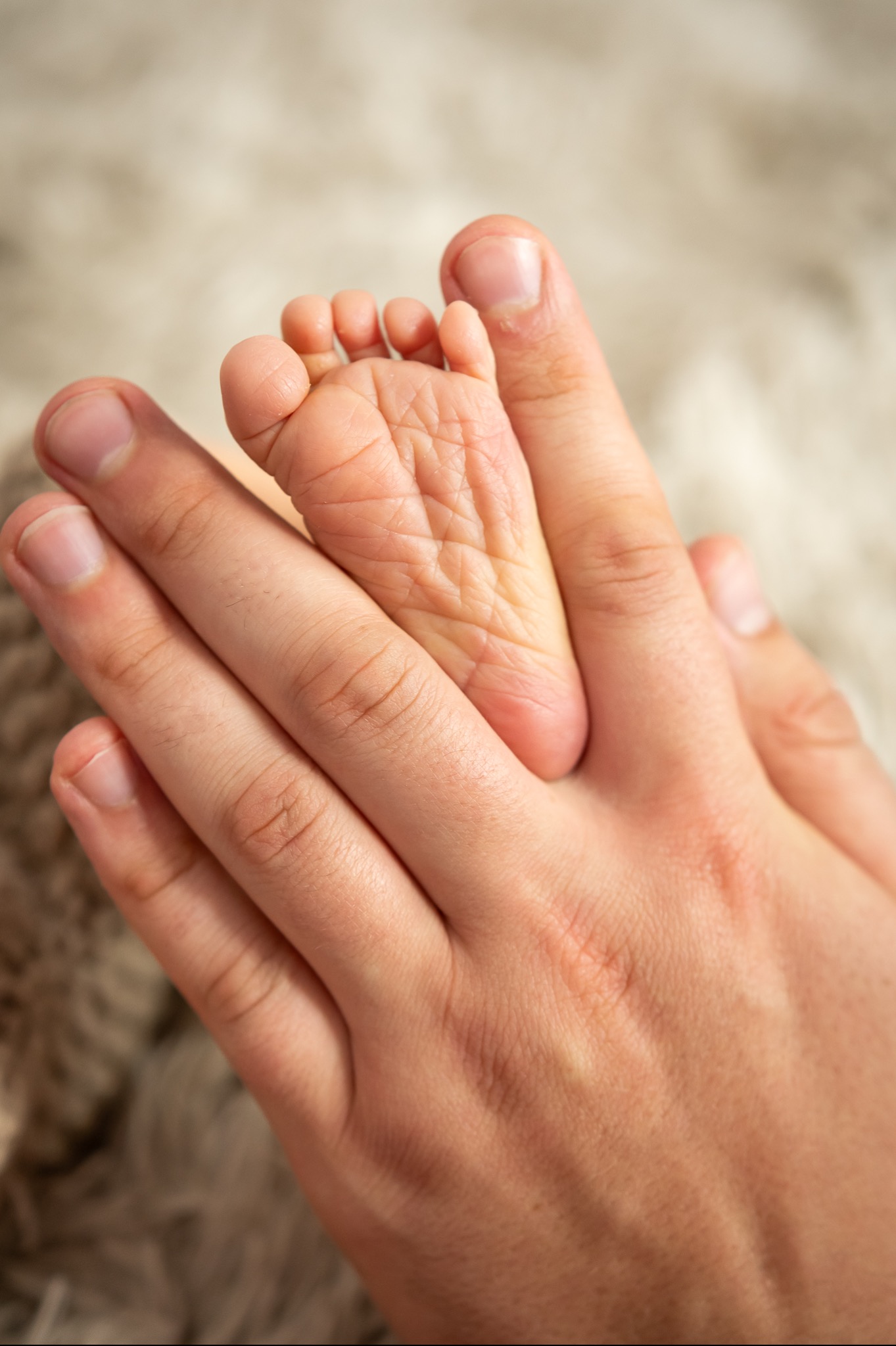 Dłoń dorosłego delikatnie trzyma drobną stopę noworodka na puszystym, beżowym tle.