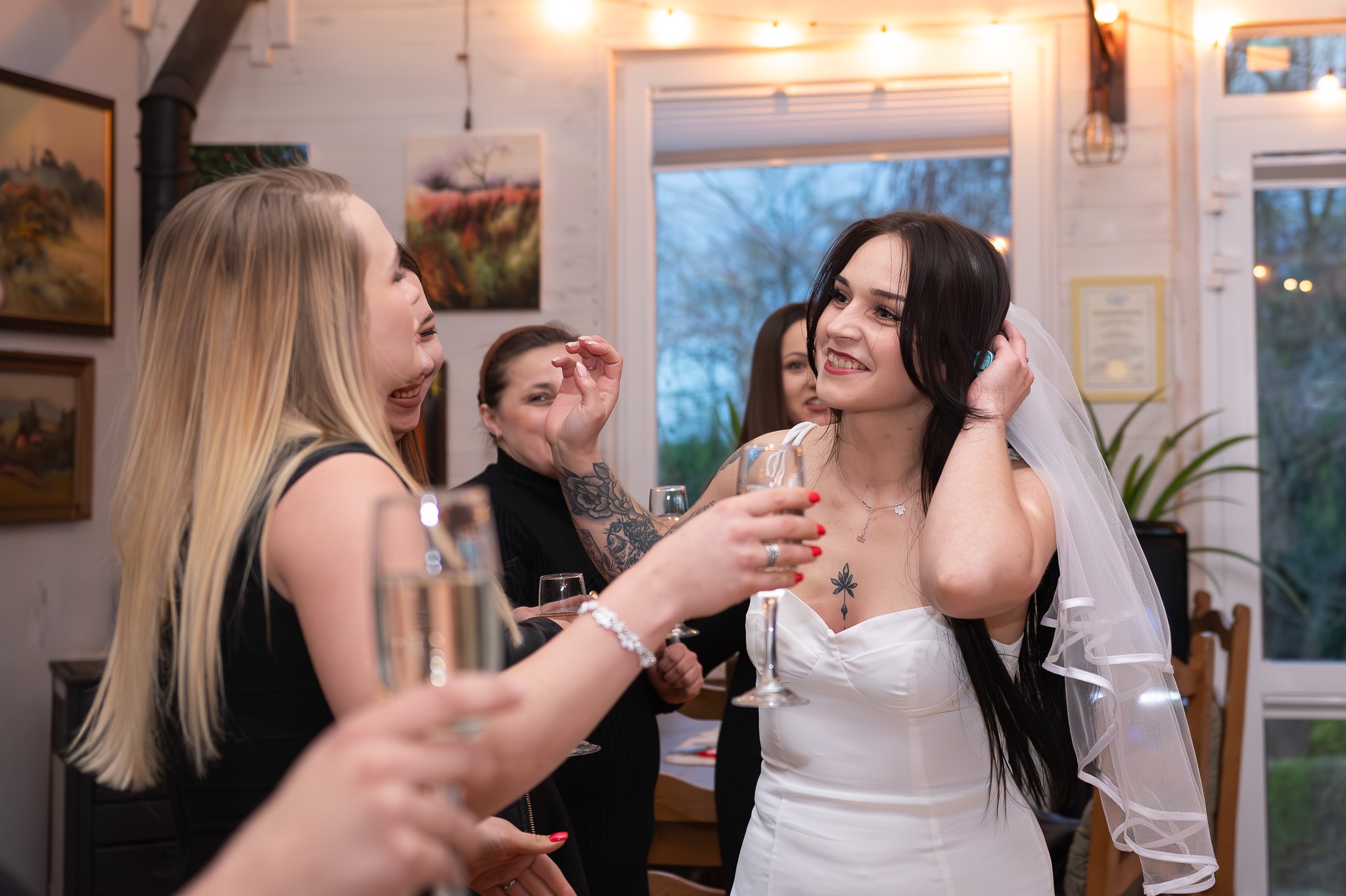 Uśmiechnięta panna młoda w białej sukni i welonie, wznosi toast z przyjaciółkami w jasnym pomieszczeniu z dekoracyjnym oświetleniem i obrazami na ścianie. Widoczne tatuaże na ramieniu jednej z kobiet.