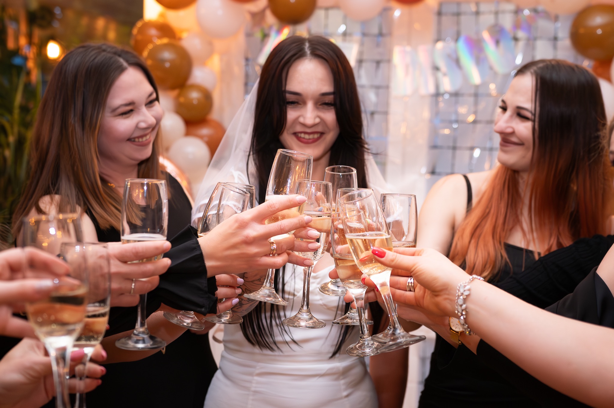 Panna młoda w welonie i dwie kobiety wznoszą toast szampanem w kieliszkach na tle dekoracji z balonów i błyszczącej kurtyny.
