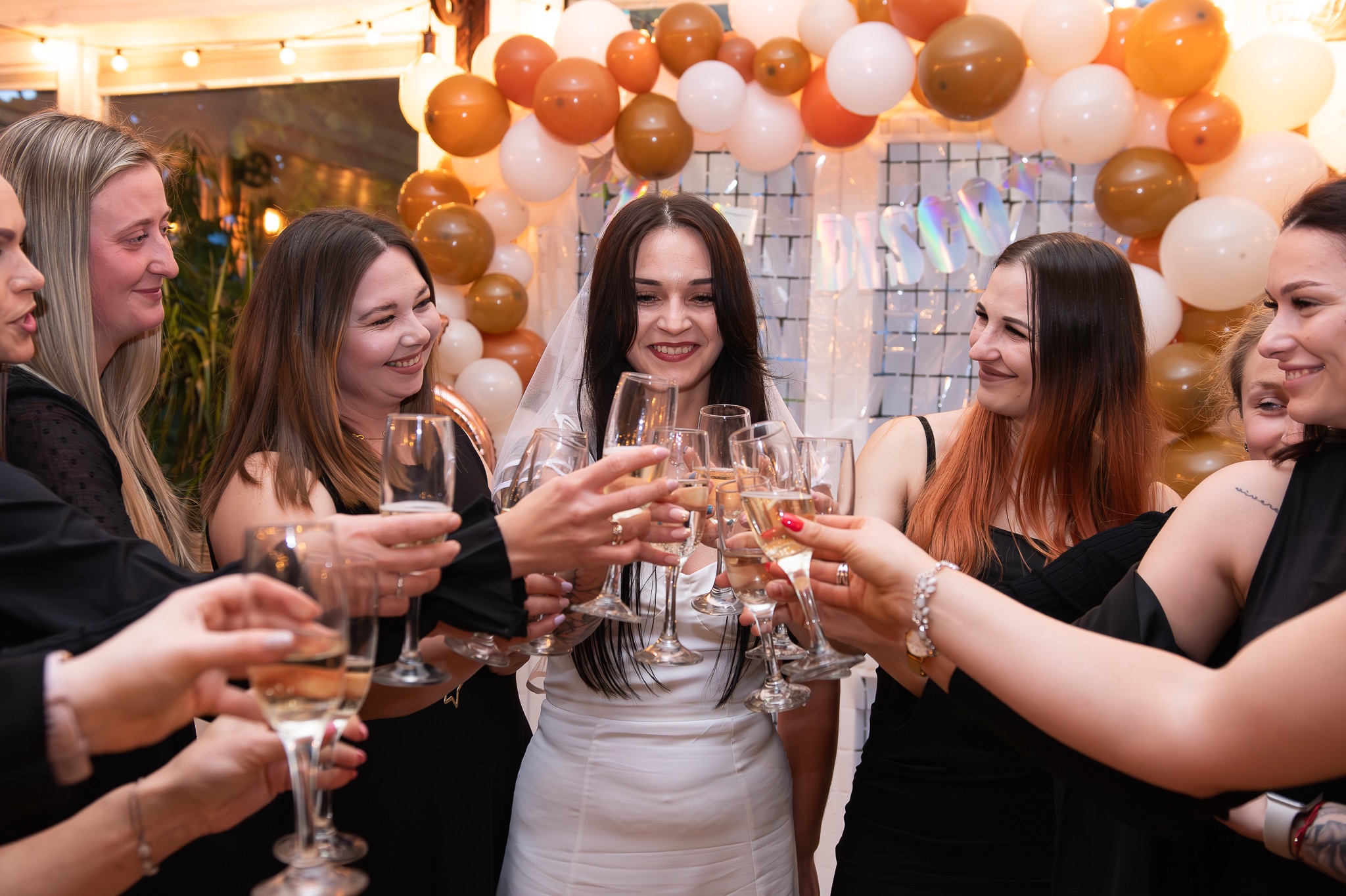 Uśmiechnięta panna młoda w białej sukni i welonie, otoczona druhnami w czarnych sukienkach, wznosi toast szampanem na tle dekoracji z białych i brązowych balonów.