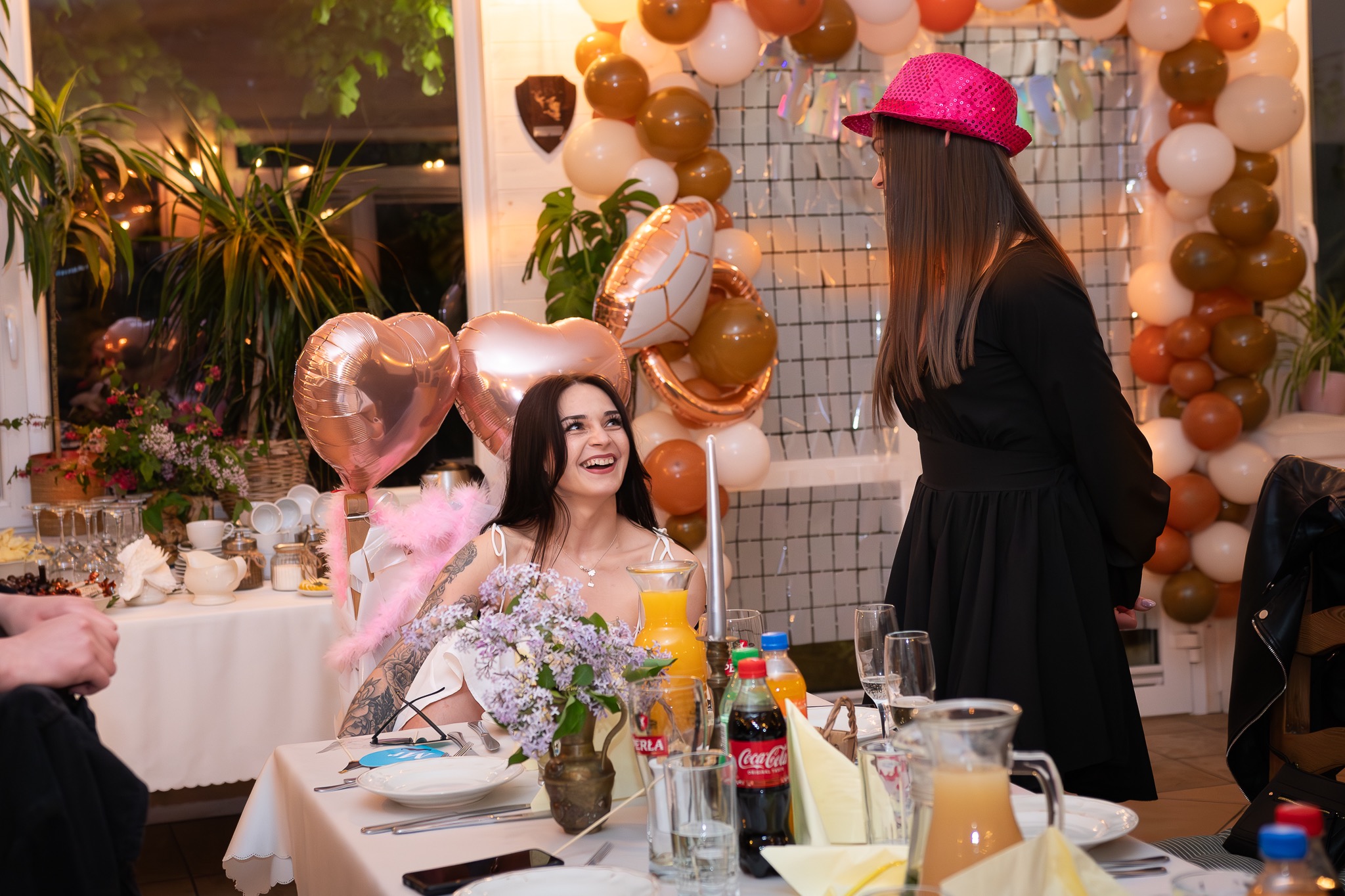 Uśmiechnięta brunetka siedzi przy stole z bukietem bzu, balonami w kształcie serc i napojami, rozmawiając z kobietą w różowym kapeluszu na tle dekoracji z balonów.