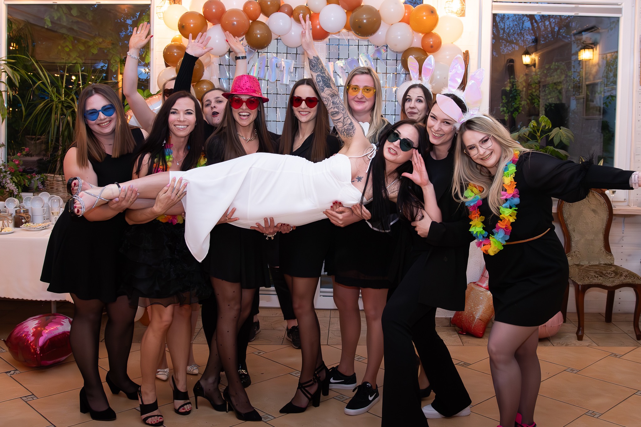 Panna młoda w białej sukience unoszona przez druhny w czarnych strojach na tle dekoracji z balonów w kolorach ziemi podczas wieczoru panieńskiego.