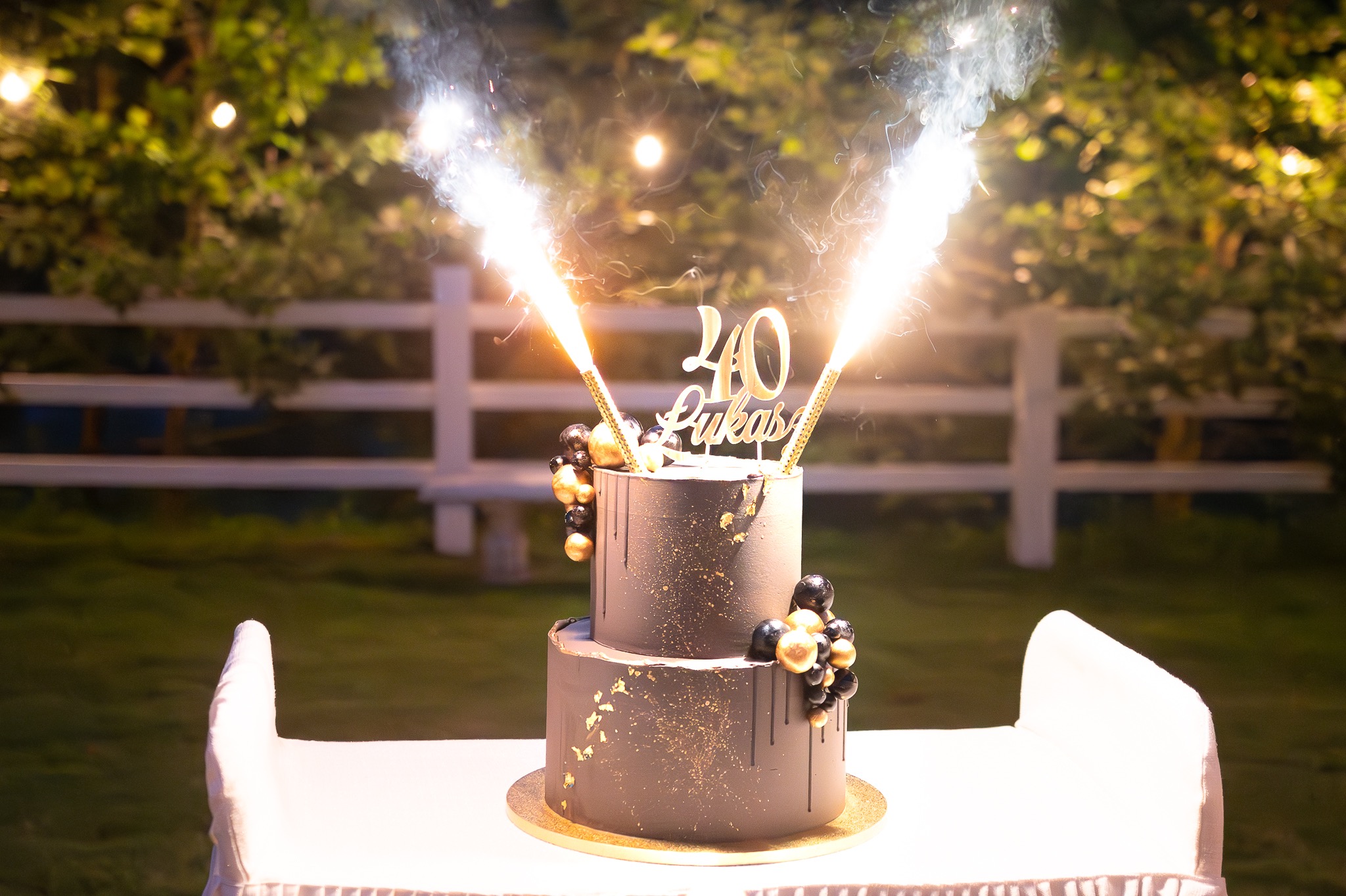 Dwupiętrowy tort urodzinowy w kolorze ciemnoszarym, ozdobiony złotymi akcentami, czarnymi i złotymi kulkami oraz płonącymi fontannami, z napisem '40 Lukas' na tle zieleni i białego płotu.