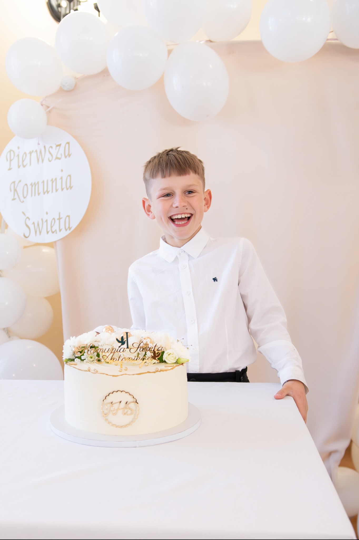 Uśmiechnięty chłopiec w białej koszuli pozuje za tortem komunijnym z napisem i dekoracją kwiatową, w tle białe balony i dekoracja z napisem 'Pierwsza Komunia Święta'.