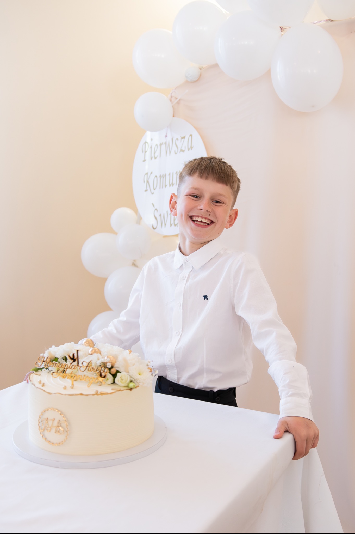 Uśmiechnięty chłopiec w białej koszuli pozuje za tortem komunijnym udekorowanym białymi kwiatami i złotymi akcentami, w tle białe balony i dekoracja z napisem 'Pierwsza Komunia Święta'.