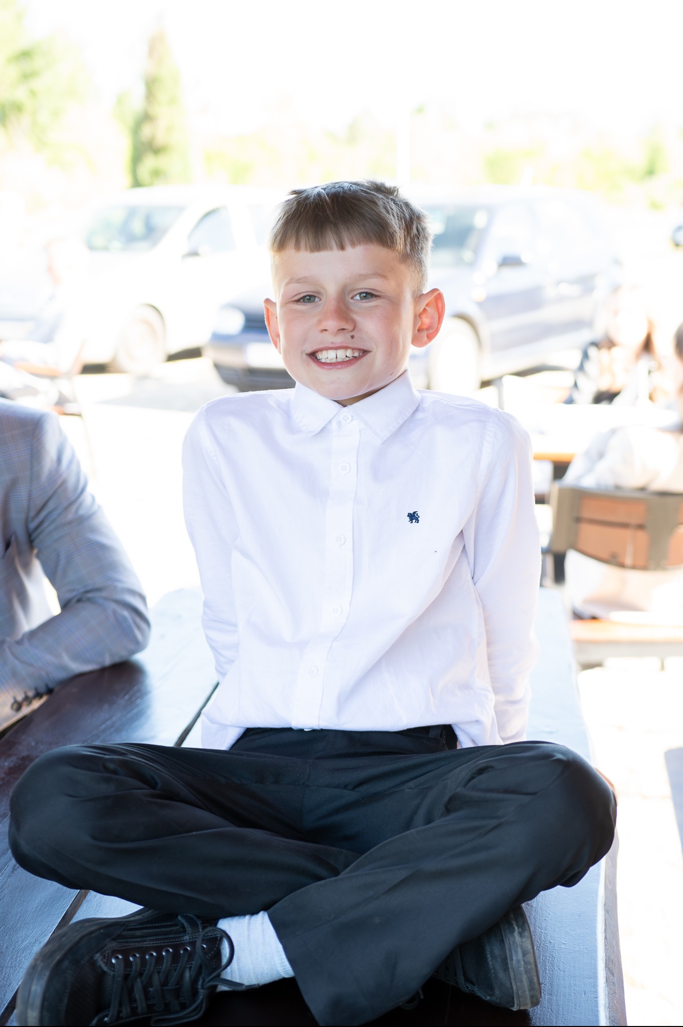 Uśmiechnięty chłopiec w białej koszuli i czarnych spodniach siedzi po turecku na drewnianym stole, w tle zaparkowane samochody i osoby.