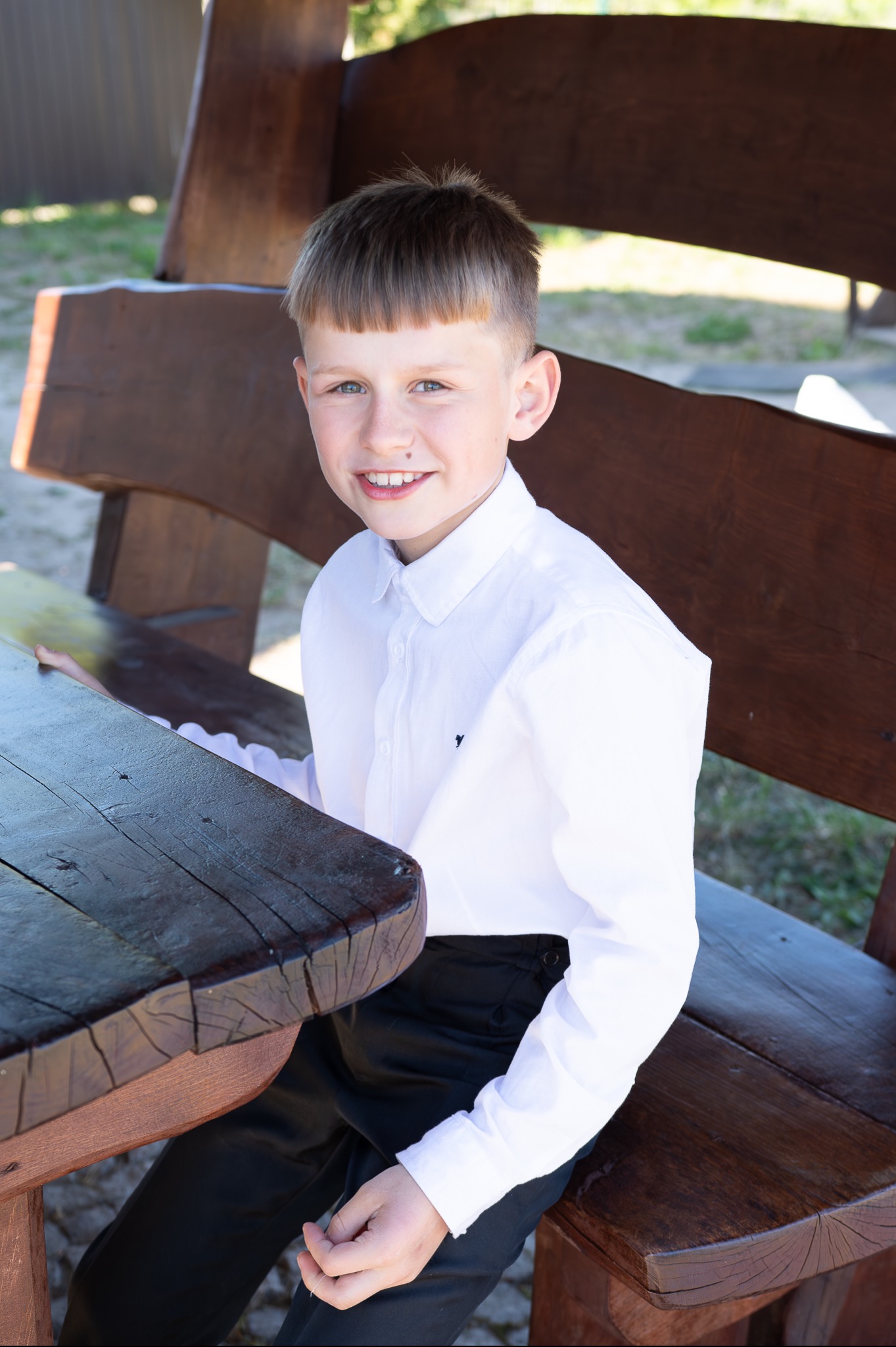 Uśmiechnięty chłopiec w białej koszuli siedzi przy drewnianym stole na tle drewnianej ławki i zieleni. Zdjęcie portretowe w naturalnym oświetleniu.