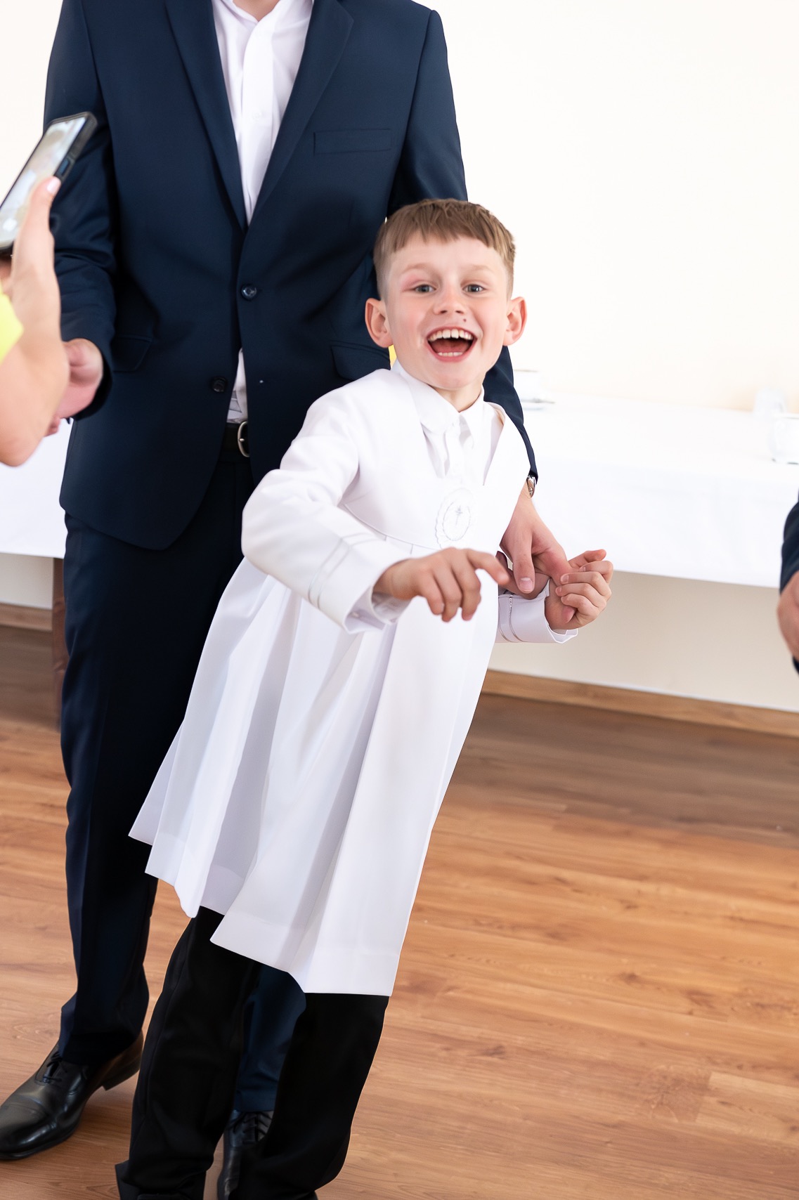 Uśmiechnięty chłopiec w albie komunijnej, prowadzony przez mężczyznę w garniturze, uchwycony w ruchu w Ostrowcu Świętokrzyskim. W tle widoczne fragmenty stołu i inni uczestnicy uroczystości.