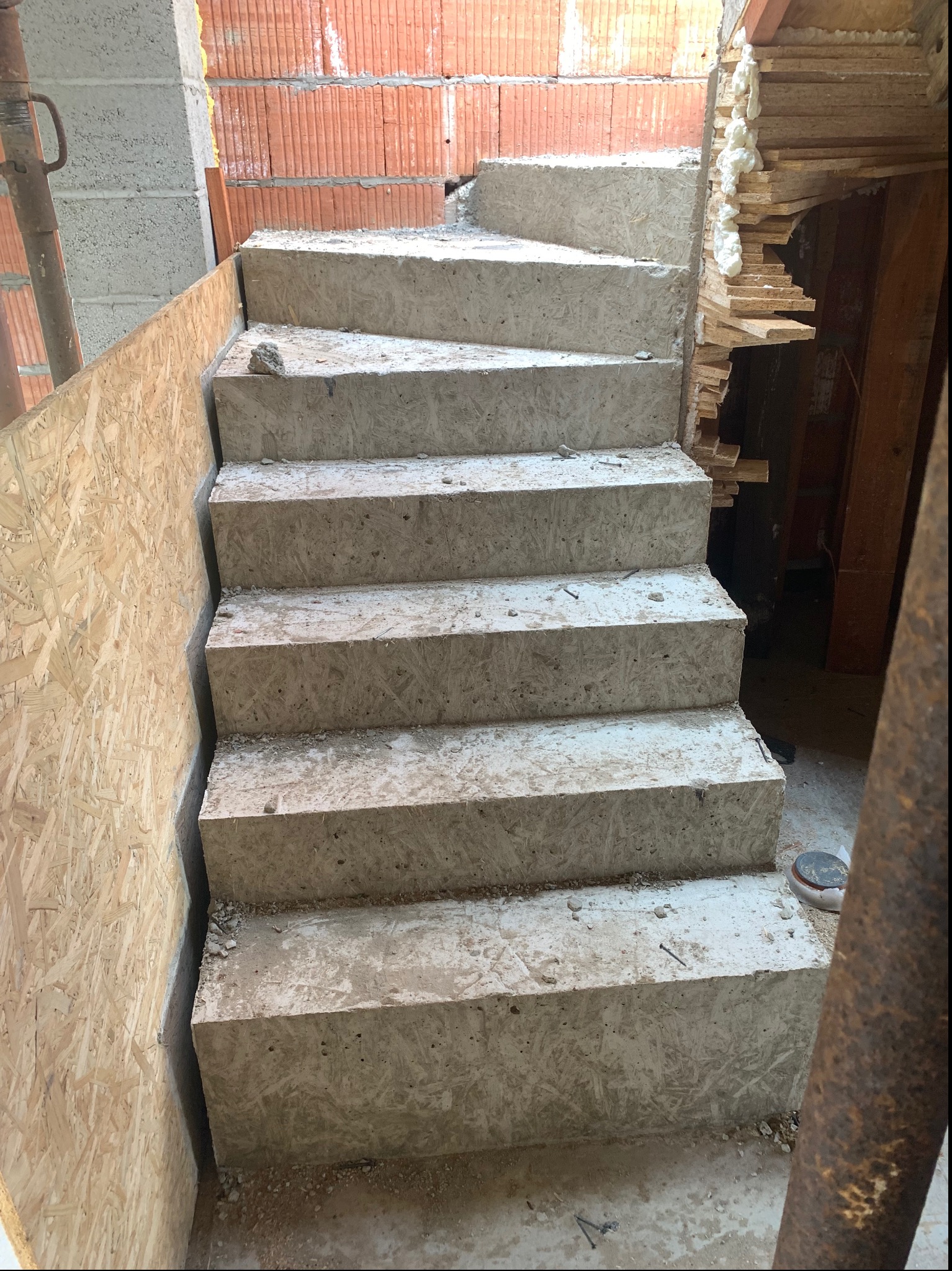 Betonowe schody tymczasowe w surowym stanie, widoczne ślady szalunku i niedoskonałości betonu, osadzone pomiędzy ścianą z cegły dziurawki a płytą OSB.