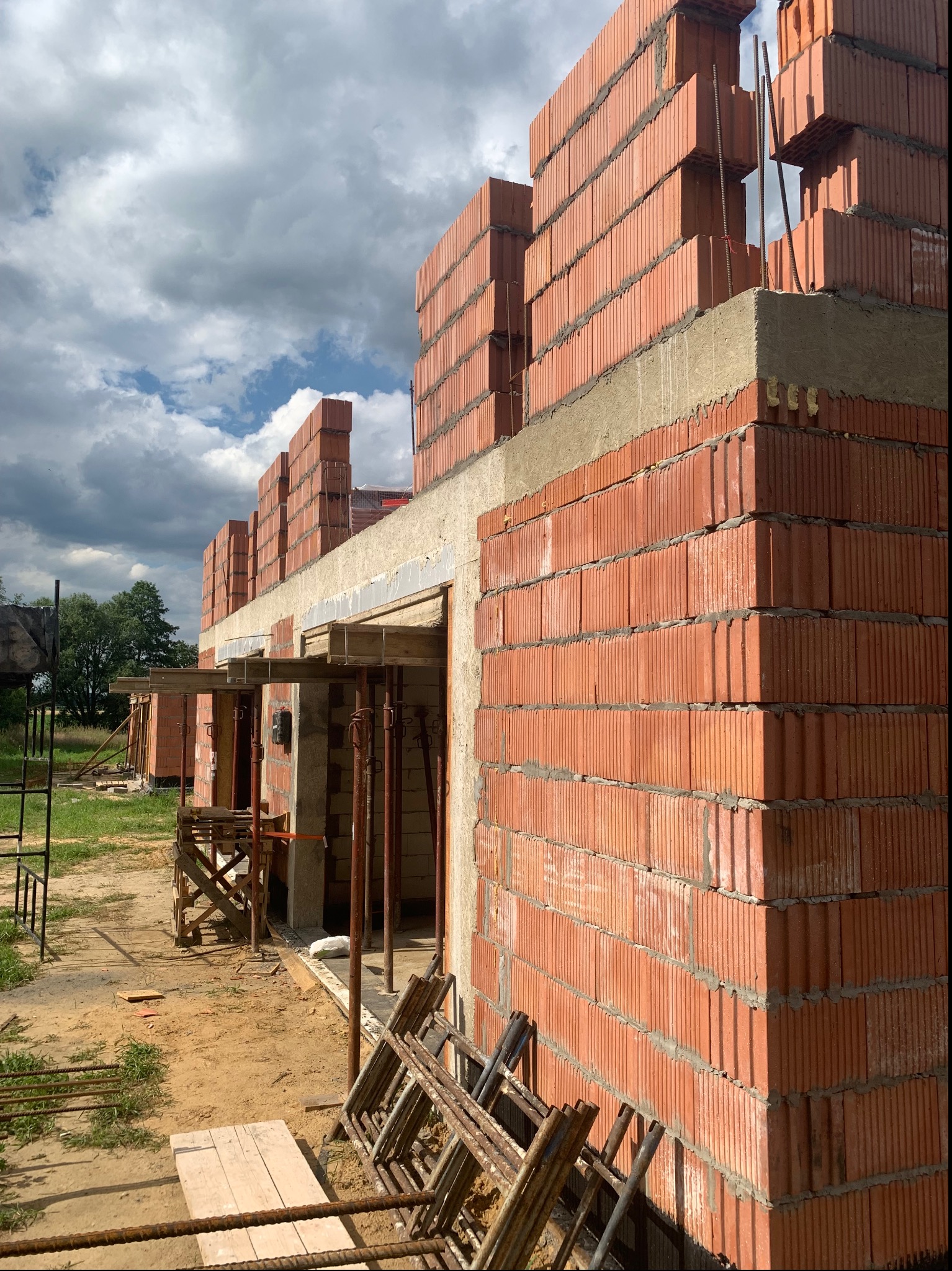 Wznoszenie ścian zewnętrznych budynku z czerwonej cegły ceramicznej, widoczne zbrojenia i betonowe nadproża, rusztowania i drewniane podpory w tle, pochmurne niebo.