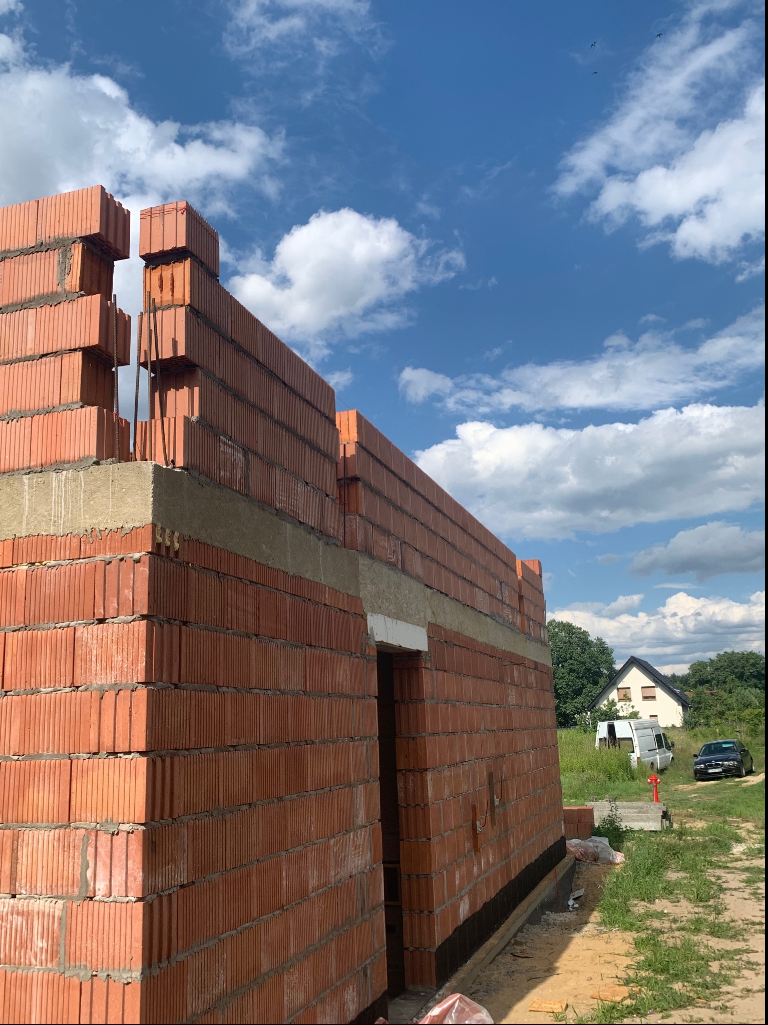 Wznoszenie ściany budynku z czerwonej cegły ceramicznej Porotherm, widoczne zbrojenie pionowe i poziome, w tle błękitne niebo z białymi chmurami oraz zabudowa jednorodzinna.