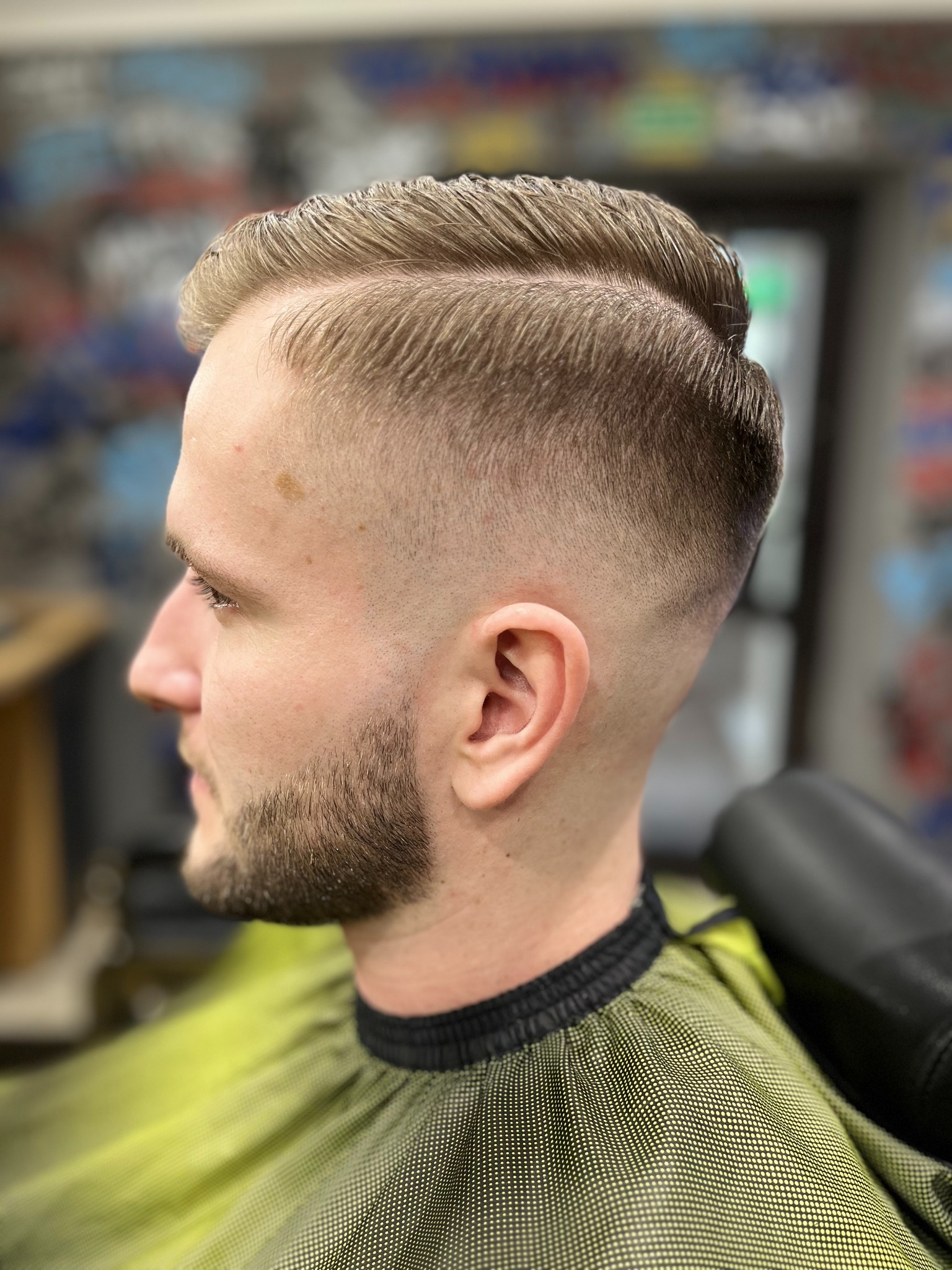 Męska fryzura z przedziałkiem na bok, cieniowane boki i krótka broda, widoczne z profilu, klient ubrany w pelerynę fryzjerską.