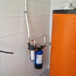 Nowa instalacja filtrów wody z rurami na tle białych płytek i pomarańczowego bojlera. Widoczne zawory, złączki i elementy mocujące rury do ściany.