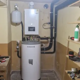 Biały, cylindryczny bojler De Dietrich z czarnymi rurami w kotłowni, obok półka z narzędziami. Widoczne elementy instalacji grzewczej i zbiornik wyrównawczy.