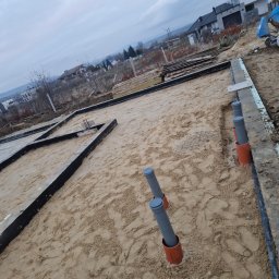 Artur Zatorski - Przygotowanie fundamentów domu: szalunki, izolacja, rury kanalizacyjne wystające z piasku. Widok z góry na plac budowy w pochmurny dzień.