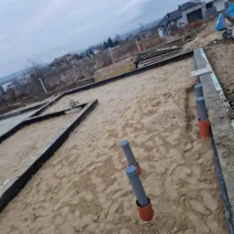 Przygotowanie fundamentów domu: szalunki, izolacja, rury kanalizacyjne wystające z piasku. Widok z góry na plac budowy w pochmurny dzień.