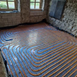 Pomarańczowe rury ogrzewania podłogowego ułożone w pętle na folii, widoczne na tle surowych, kamiennych ścian w starym domu z oknem. Instalacja przed wylaniem posadzki.