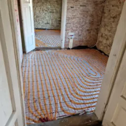 Pomarańczowe rury ogrzewania podłogowego ułożone na srebrnej folii z siatką, widoczne przez otwarte drzwi w surowym wnętrzu z ceglanymi i kamiennymi ścianami.