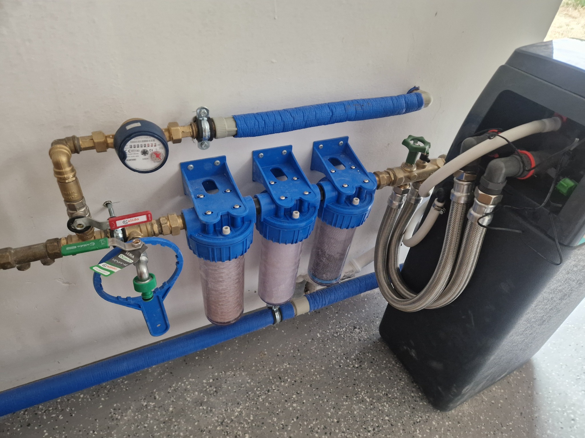 Instalacja filtrów wody z licznikiem, rurami w izolacji i zaworami. Usługa hydrauliczna w Kielcach. Widoczne trzy filtry z niebieskimi obudowami.
