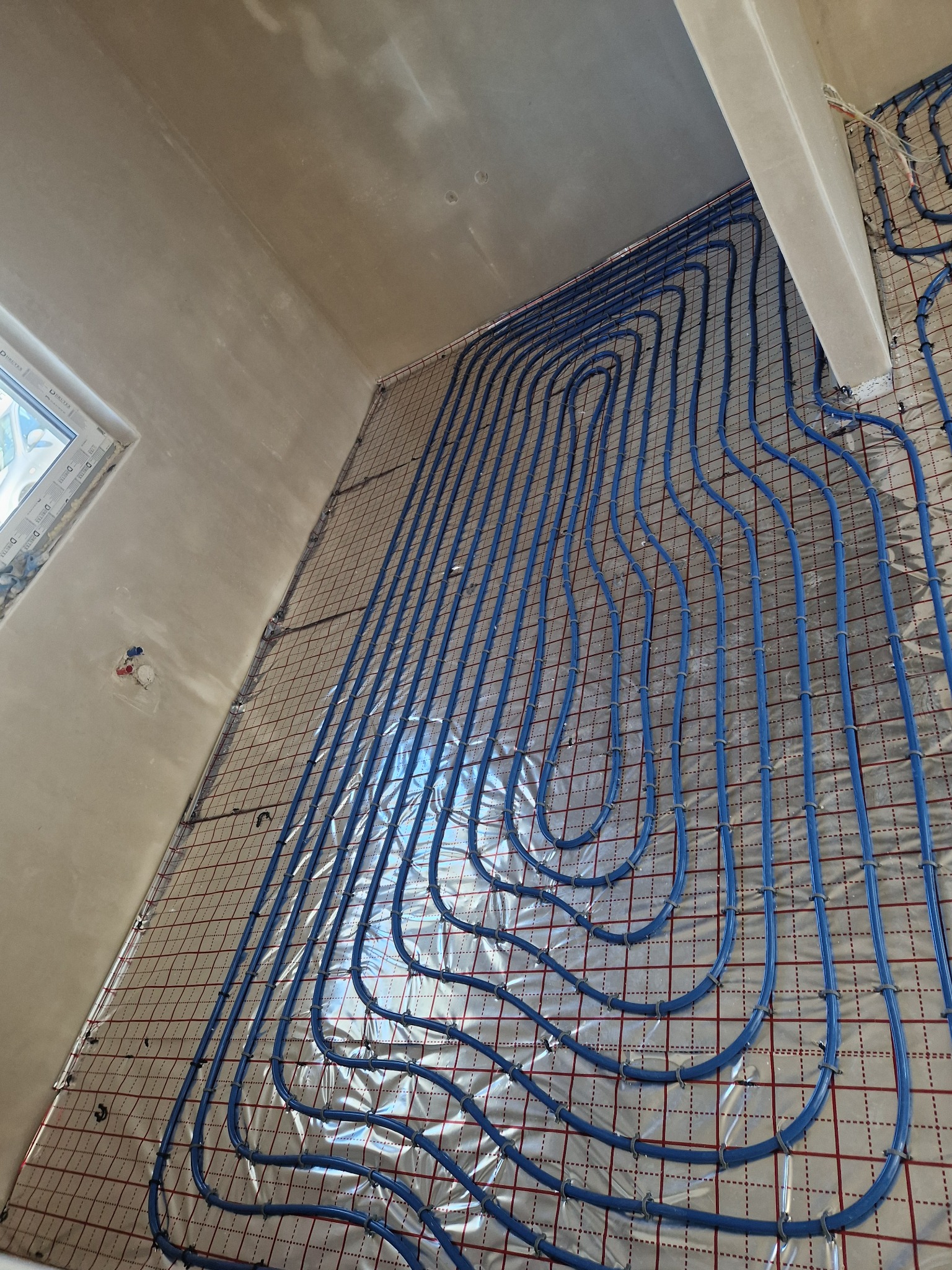 Niebieskie rury ogrzewania podłogowego ułożone na folii z siatką w surowym wnętrzu, widok z góry pod kątem.