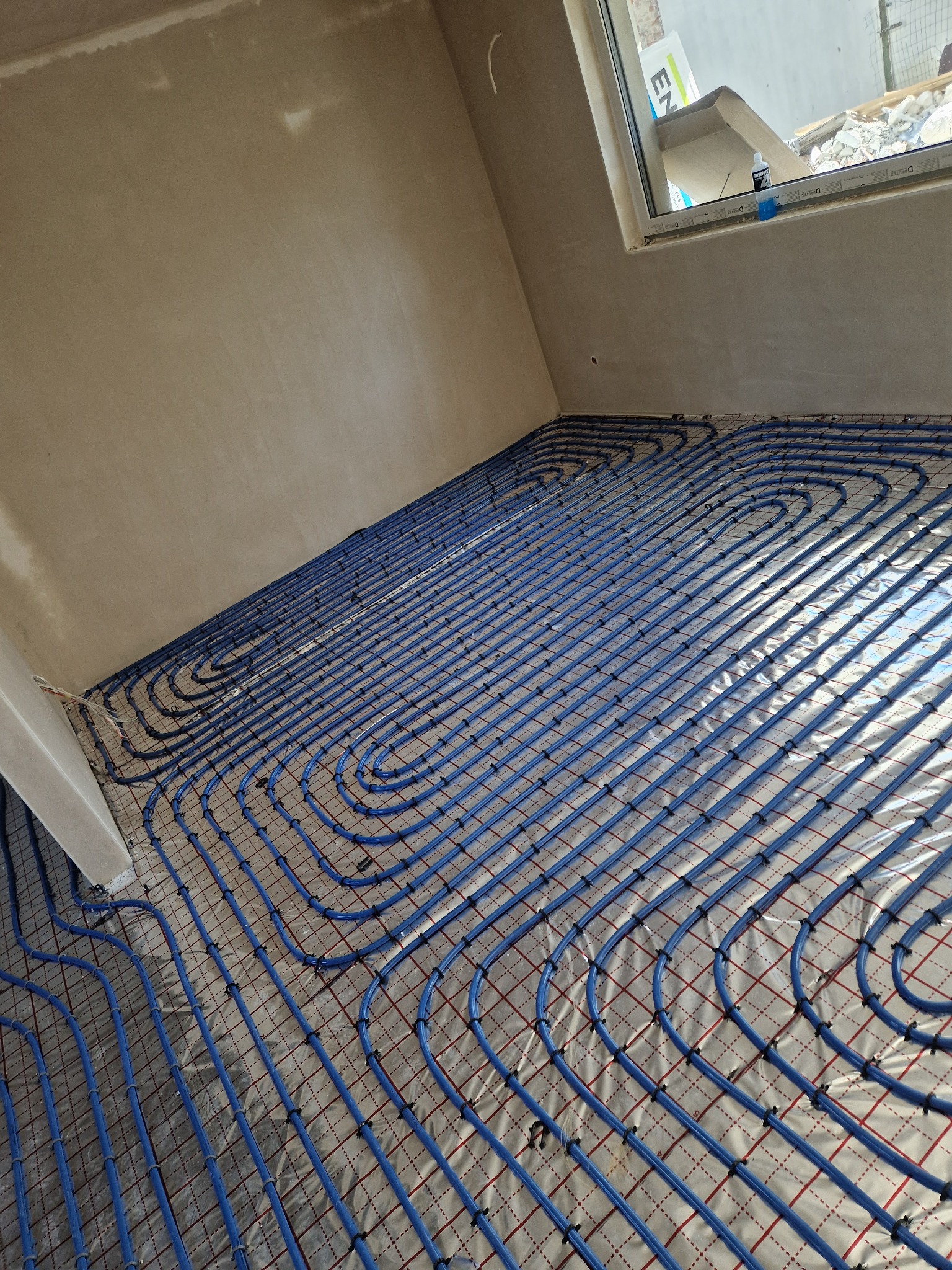 Niebieskie rury ogrzewania podłogowego ułożone spiralnie na folii izolacyjnej w surowym wnętrzu z oknem. Widoczne mocowania rur do siatki.