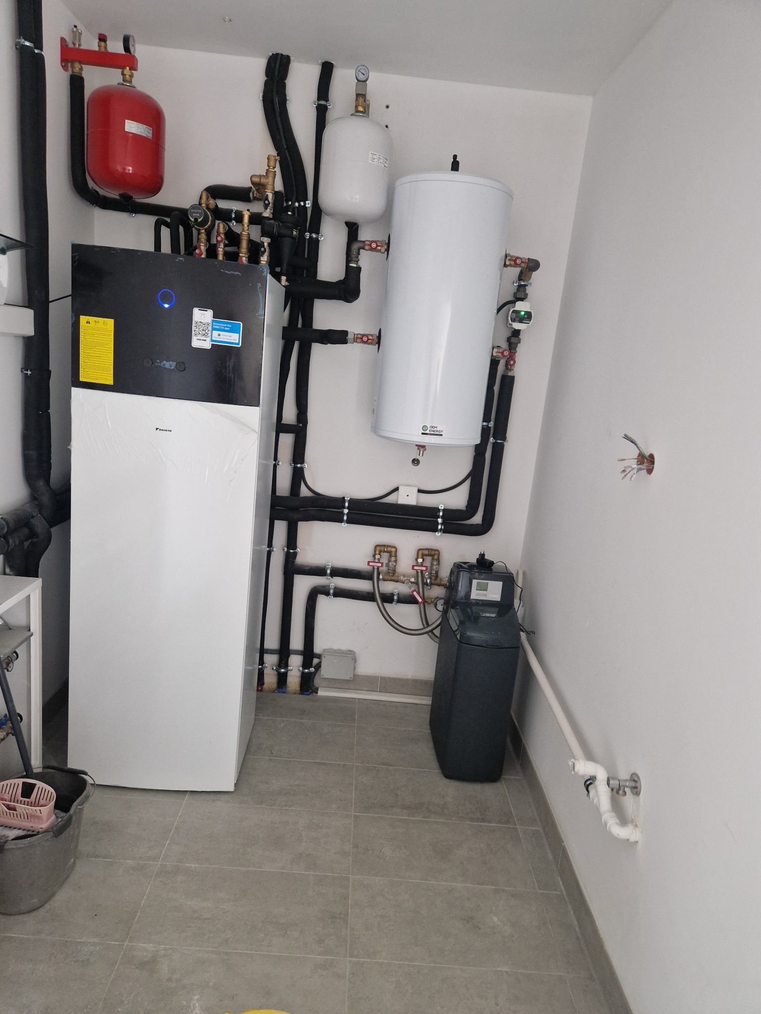 Zestaw urządzeń grzewczych: pompa ciepła Daikin, zbiornik buforowy i zmiękczacz wody, połączone rurami w pomieszczeniu kotłowni. Widoczne elementy instalacji.