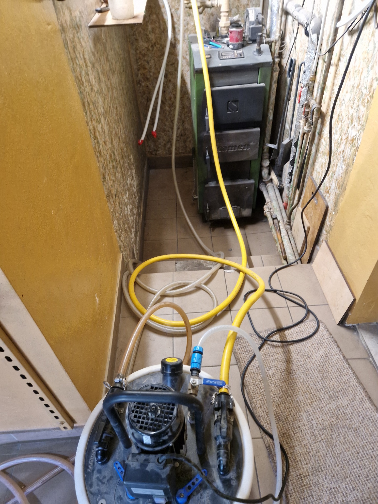 Czyszczenie kotła grzewczego z użyciem pompy i węży w pomieszczeniu technicznym. Widoczny kocioł, pompa czyszcząca, węże doprowadzające i odprowadzające wodę.