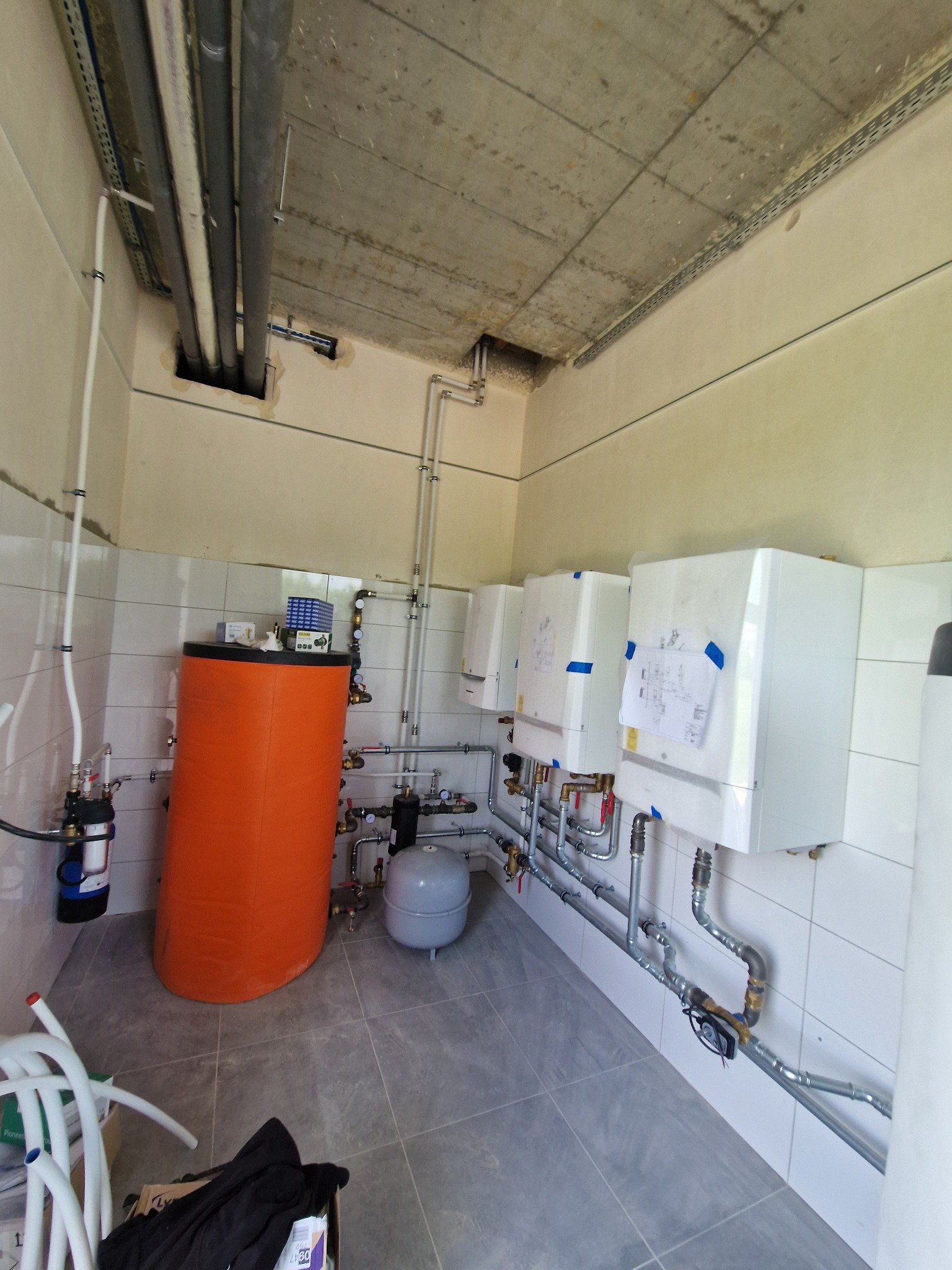 Pomieszczenie z zamontowanymi trzema białymi piecami grzewczymi, pomarańczowym zasobnikiem wody i szarą instalacją hydrauliczną na tle białych płytek.
