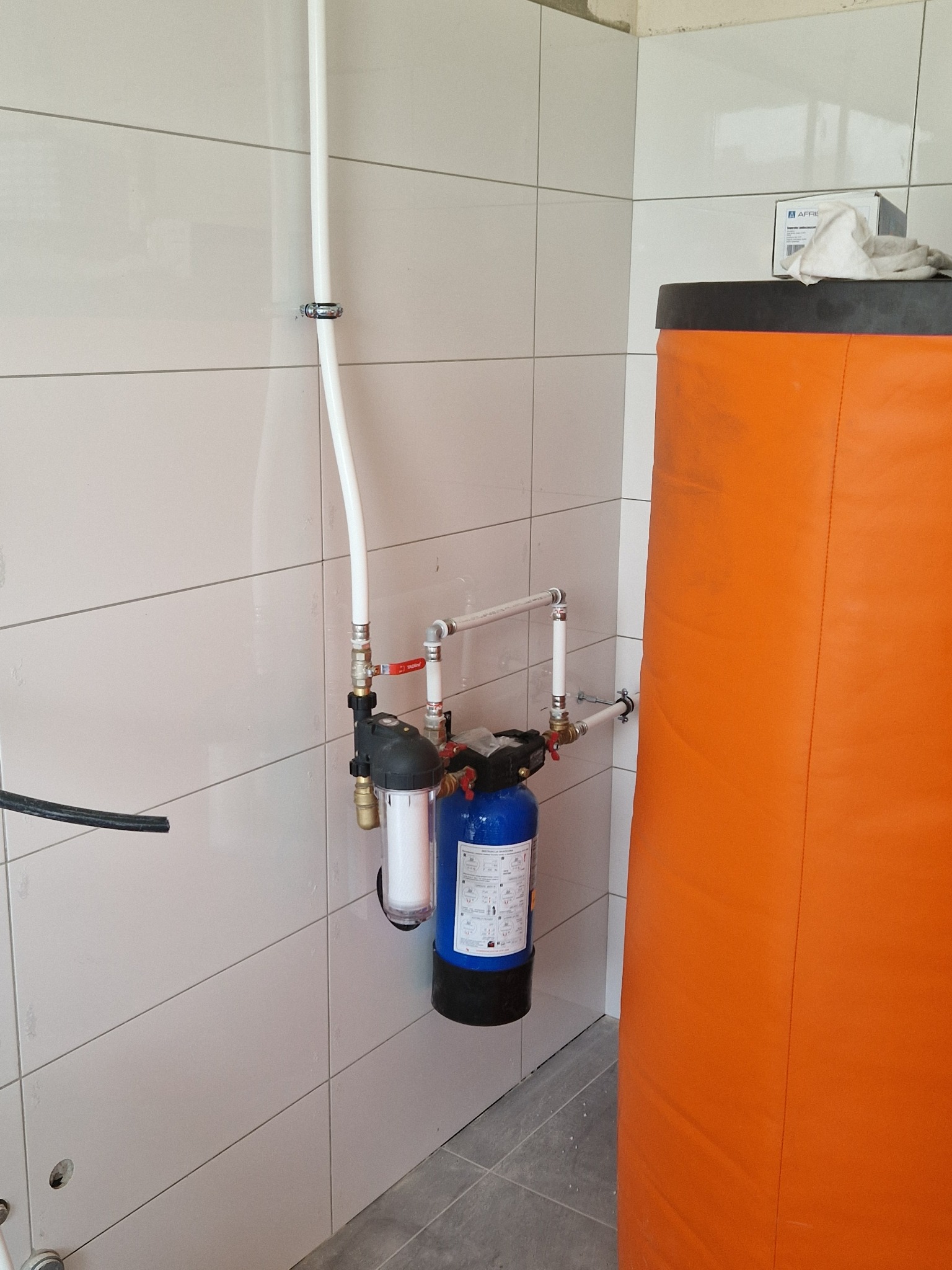 Nowa instalacja filtrów wody z rurami na tle białych płytek i pomarańczowego bojlera. Widoczne zawory, złączki i elementy mocujące rury do ściany.