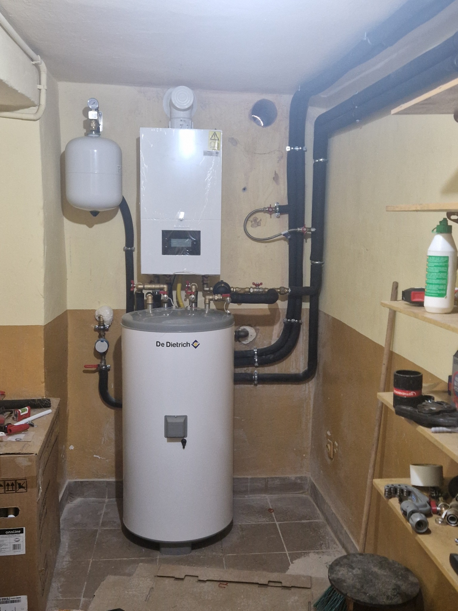 Biały, cylindryczny bojler De Dietrich z czarnymi rurami w kotłowni, obok półka z narzędziami. Widoczne elementy instalacji grzewczej i zbiornik wyrównawczy.