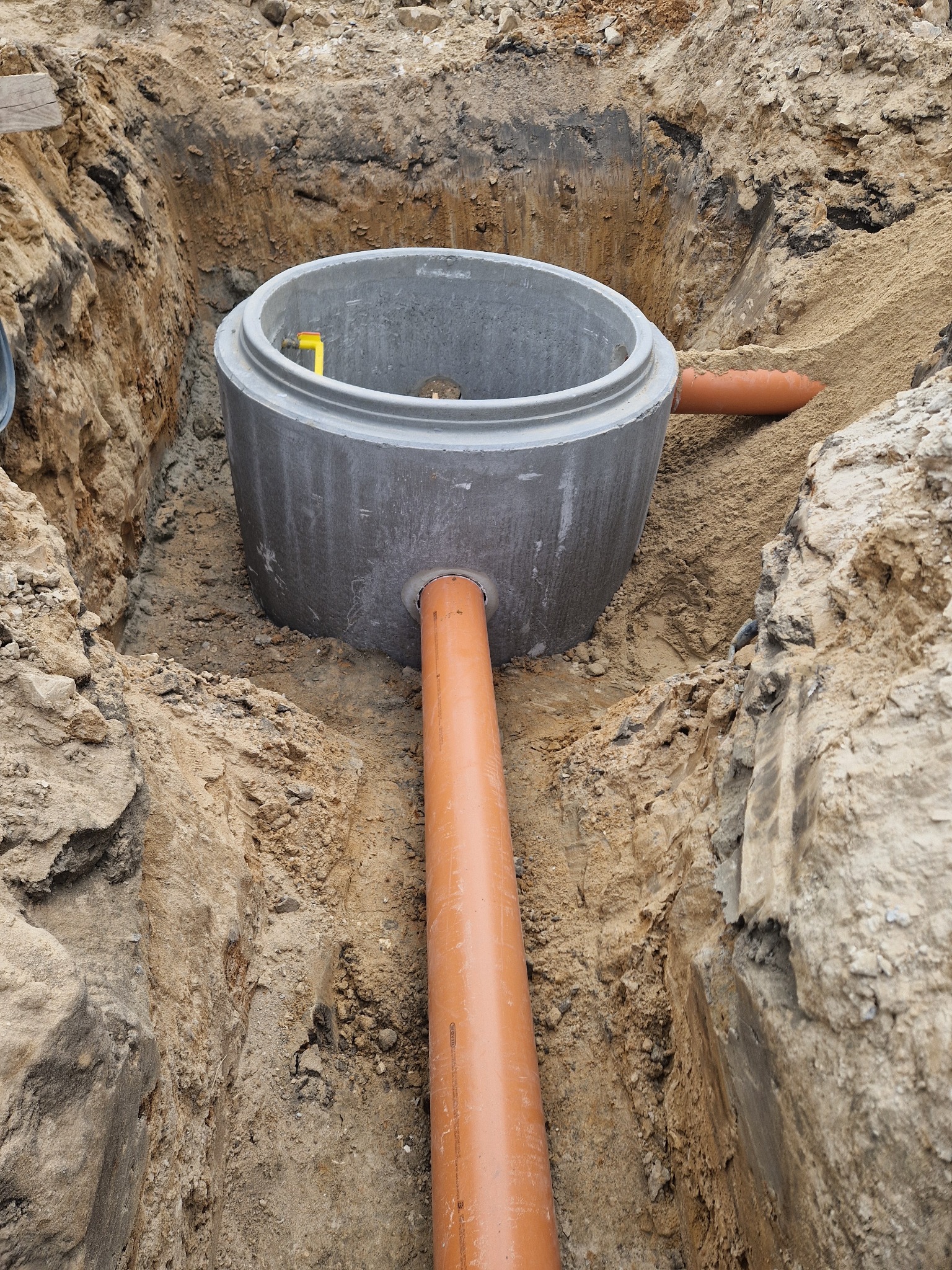 Betonowy krąg studzienny w wykopie, z podłączonymi rurami kanalizacyjnymi. Widoczne piaskowe ściany wykopu i narzędzie wewnątrz kręgu.