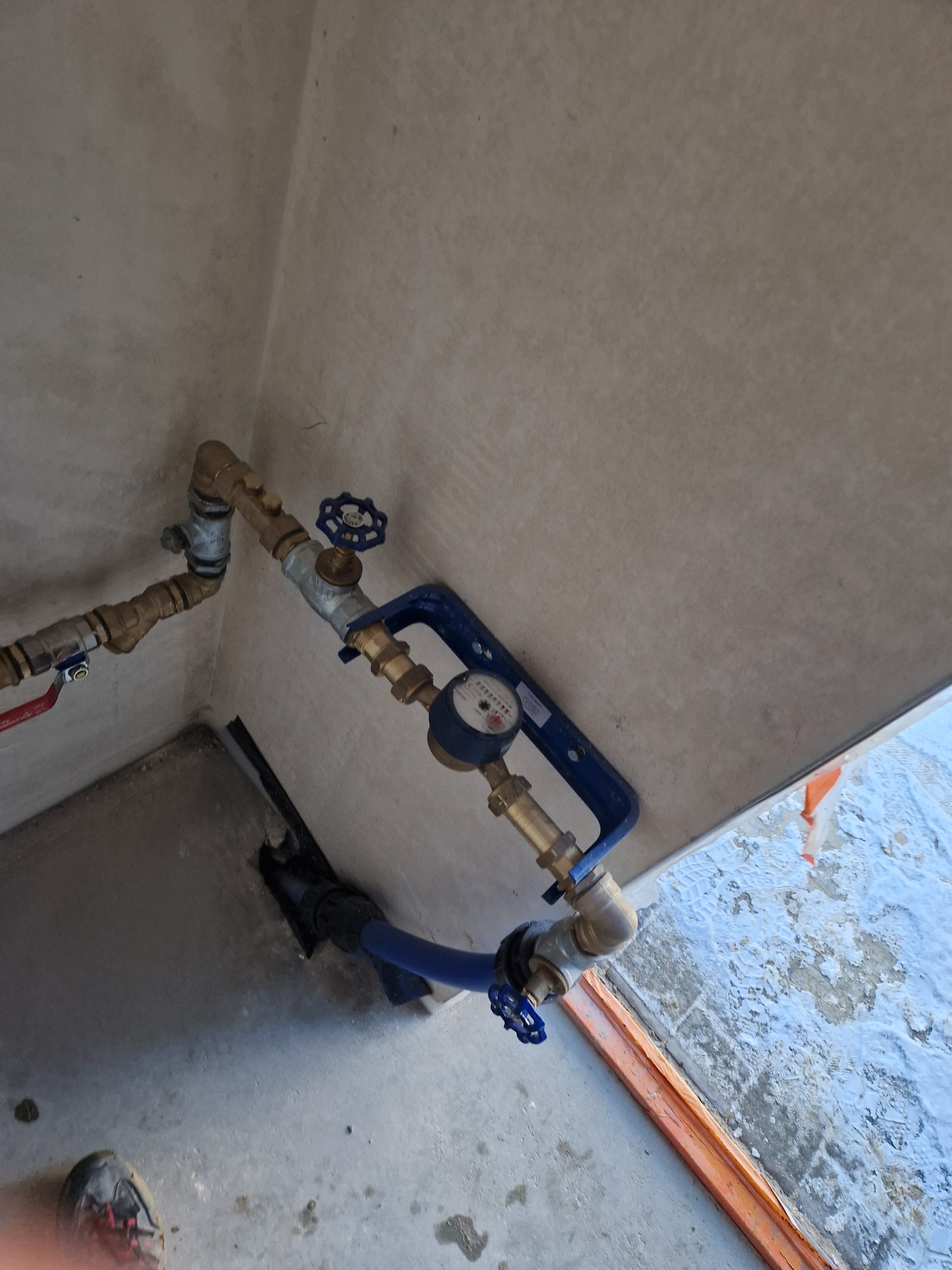 Zamontowany wodomierz z zaworami odcinającymi, połączony rurami w narożniku pomieszczenia o surowych ścianach. Widoczne elementy instalacji wodnej i fragment podłogi.