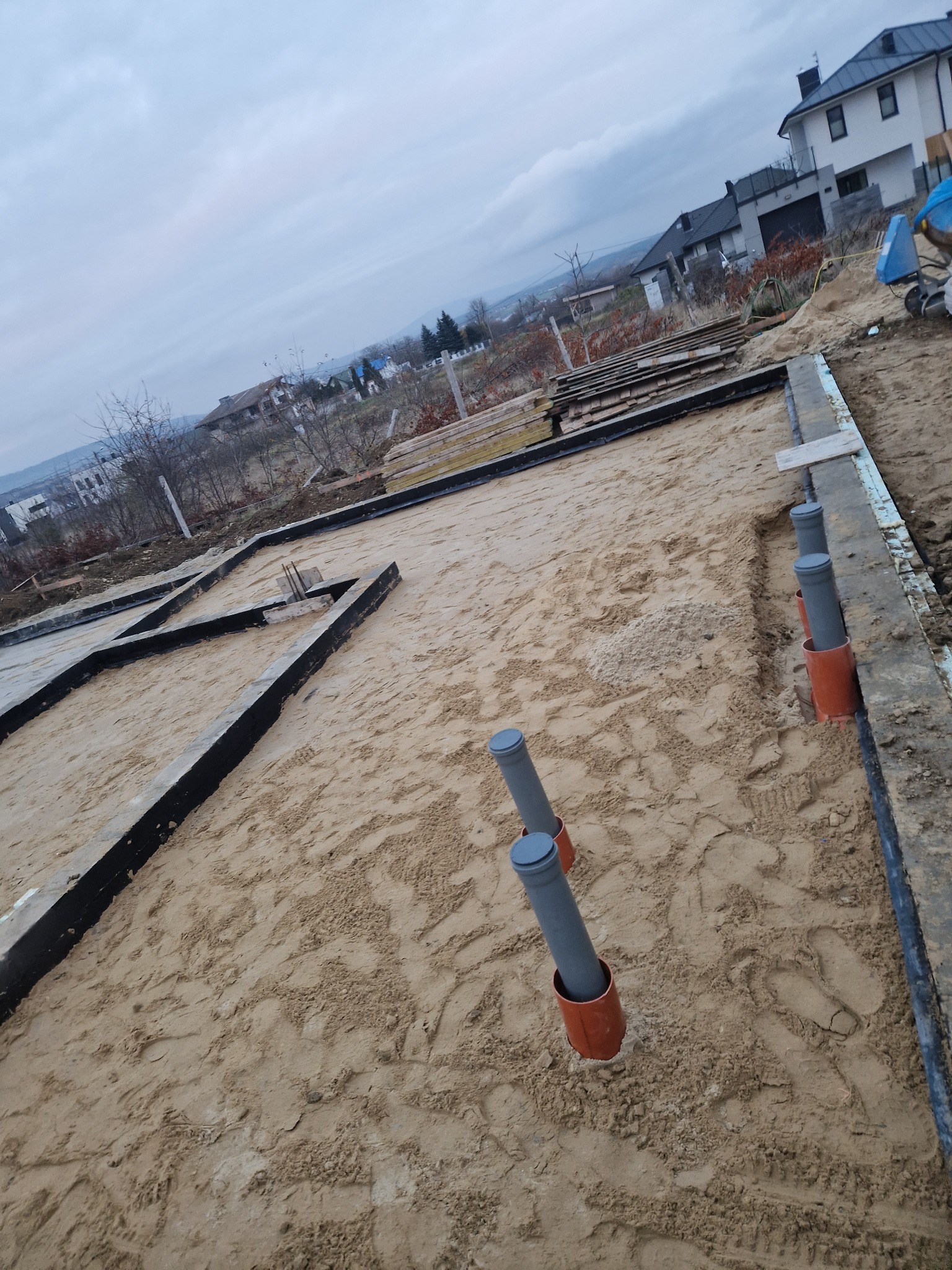 Przygotowanie fundamentów domu: szalunki, izolacja, rury kanalizacyjne wystające z piasku. Widok z góry na plac budowy w pochmurny dzień.