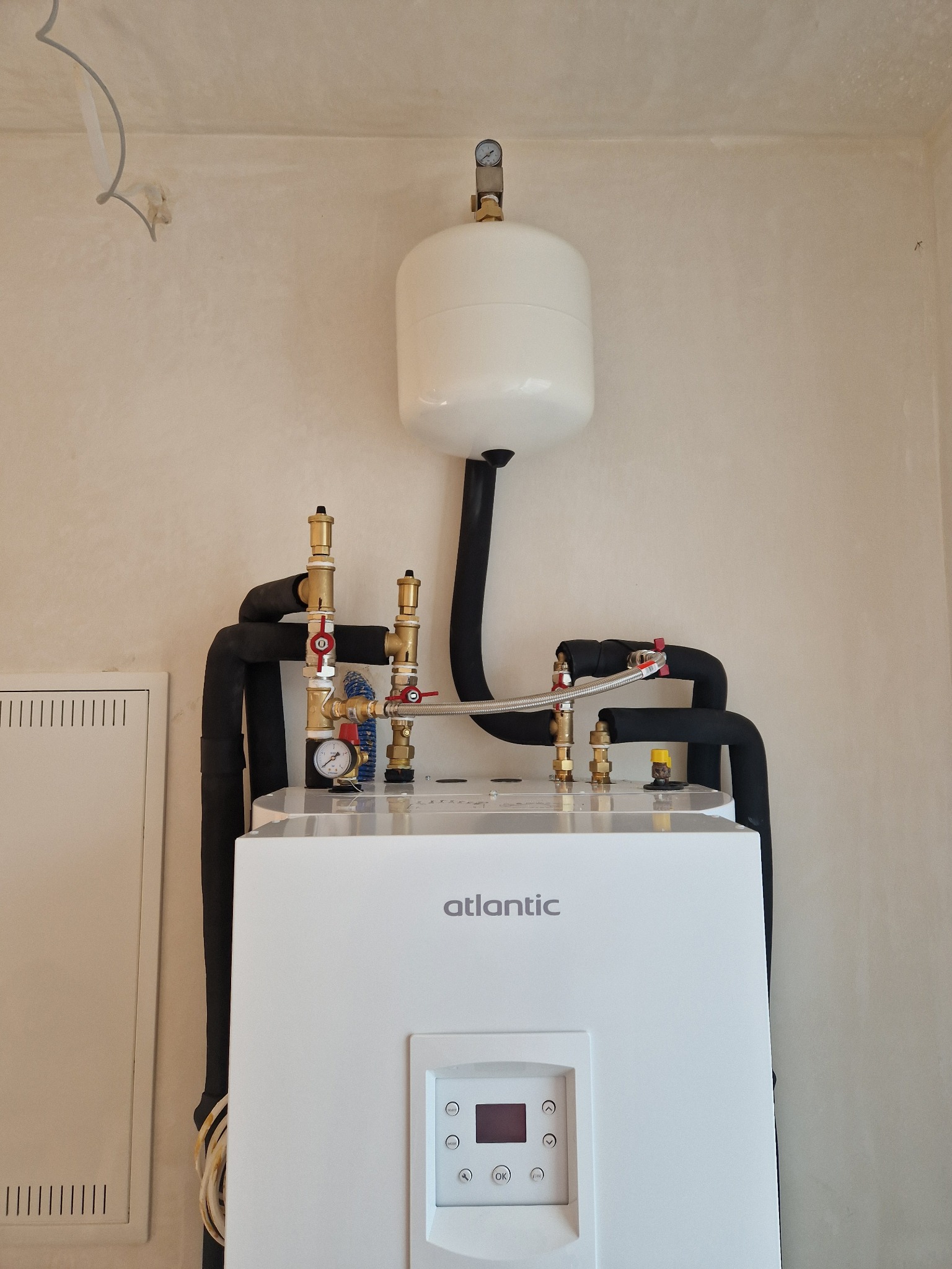 Biały piec grzewczy Atlantic z czarnymi rurami i zaworami mosiężnymi na tle beżowej ściany. Widoczny manometr i zbiornik wyrównawczy. Ujęcie od dołu.