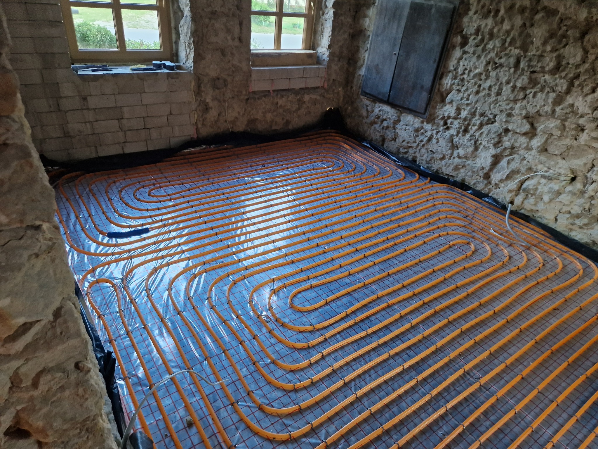 Pomarańczowe rury ogrzewania podłogowego ułożone w pętle na folii, widoczne na tle surowych, kamiennych ścian w starym domu z oknem. Instalacja przed wylaniem posadzki.