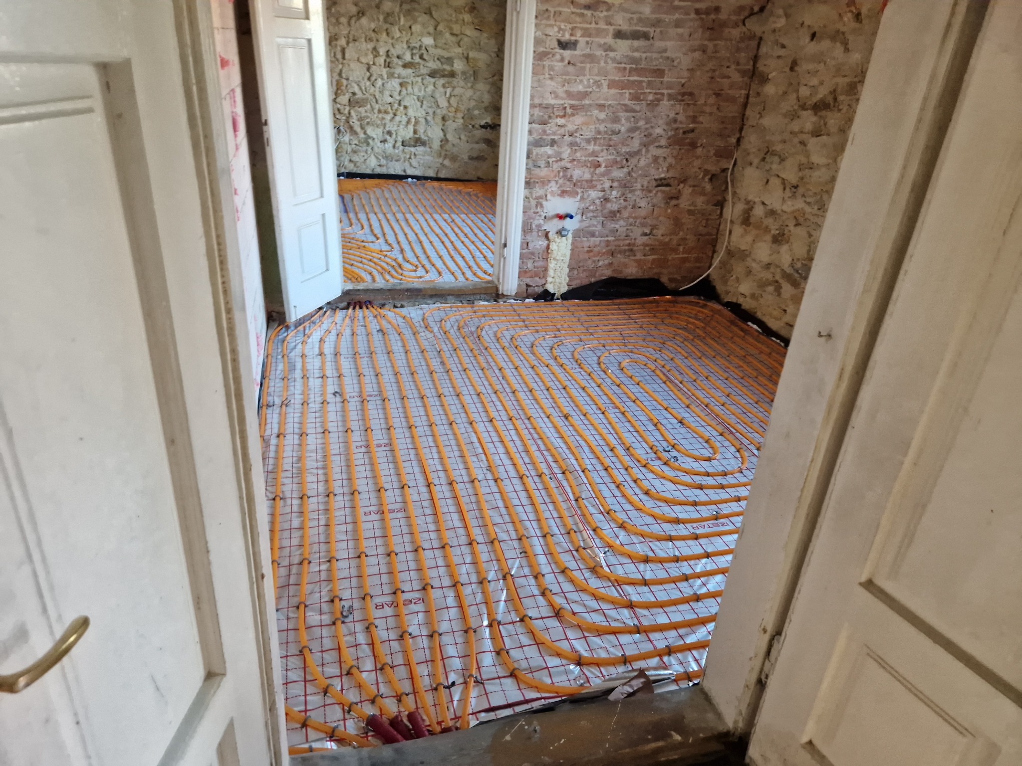 Pomarańczowe rury ogrzewania podłogowego ułożone na srebrnej folii z siatką, widoczne przez otwarte drzwi w surowym wnętrzu z ceglanymi i kamiennymi ścianami.