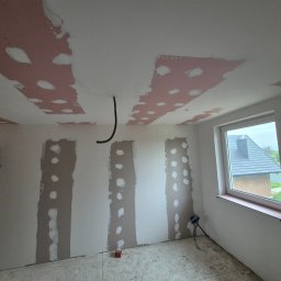 WAR-IN-BUD - Wnętrze pokoju w trakcie remontu: częściowo położona płyta karton gips na suficie i ścianach, widoczne szpachlowane łączenia, kabel zwisający z sufitu, okno z widokiem na dom.