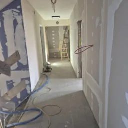 Korytarz w trakcie remontu z widocznymi ścianami z płyt kartonowo-gipsowych, szpachlowanymi łączeniami, kable wystające ze ściany, drabina i narzędzia na podłodze.