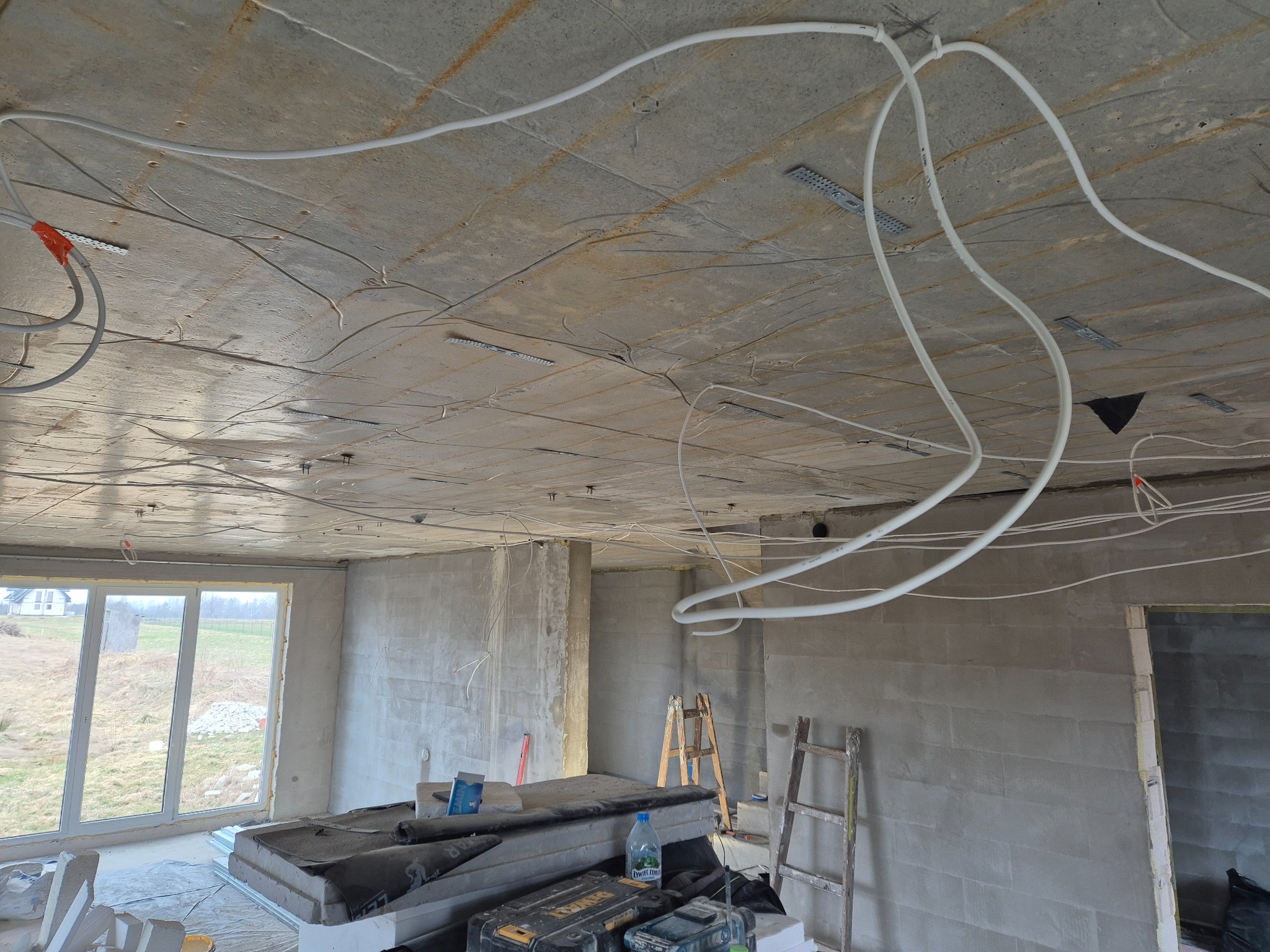 Surowy betonowy sufit z instalacją elektryczną w białych rurkach, widoczne mocowania, w tle ściany w stanie deweloperskim i okno z widokiem na krajobraz.