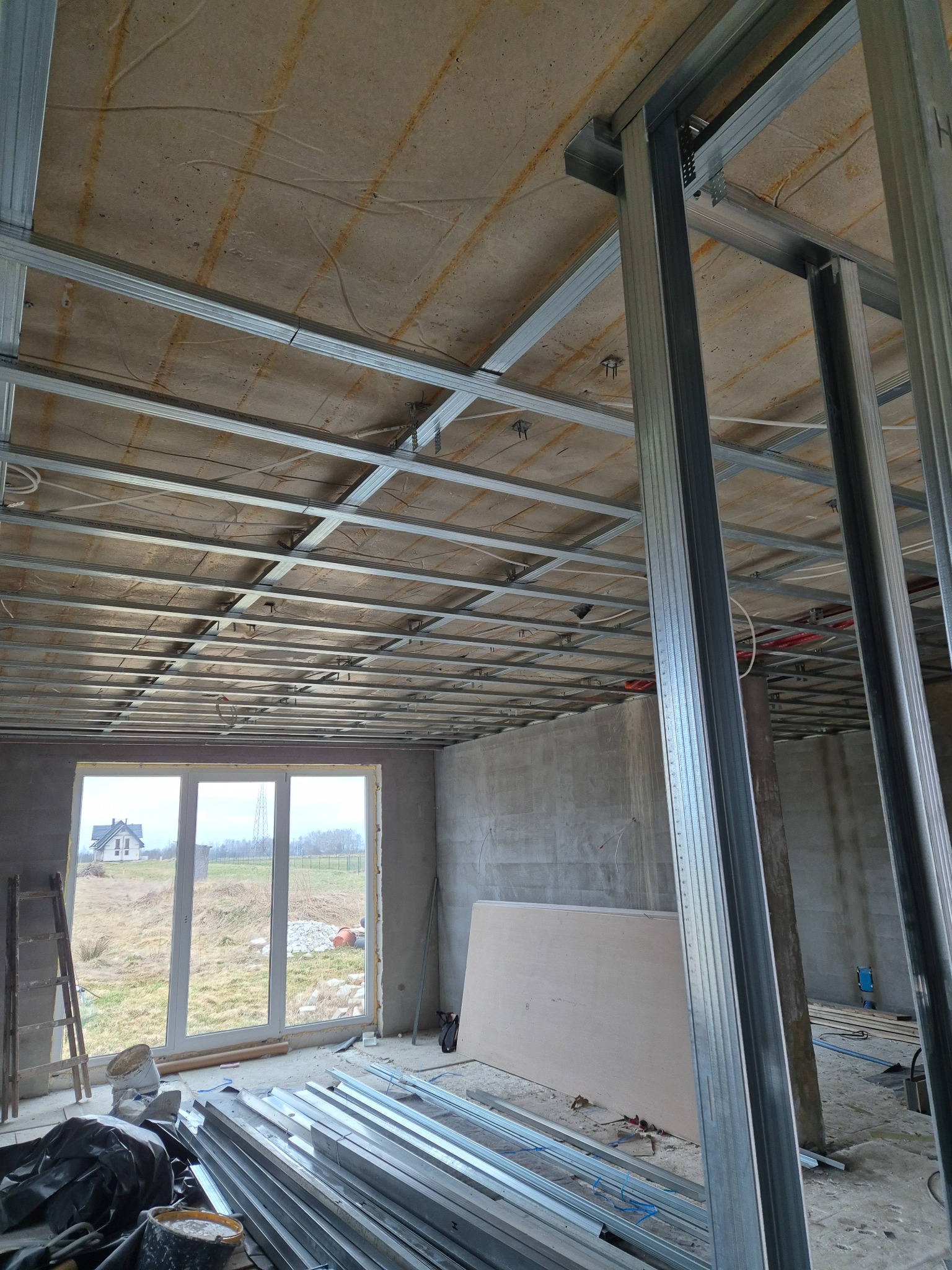 Wnętrze w trakcie montażu metalowej konstrukcji sufitu podwieszanego z widocznymi profilami, elementami instalacji i oknem na zewnątrz.