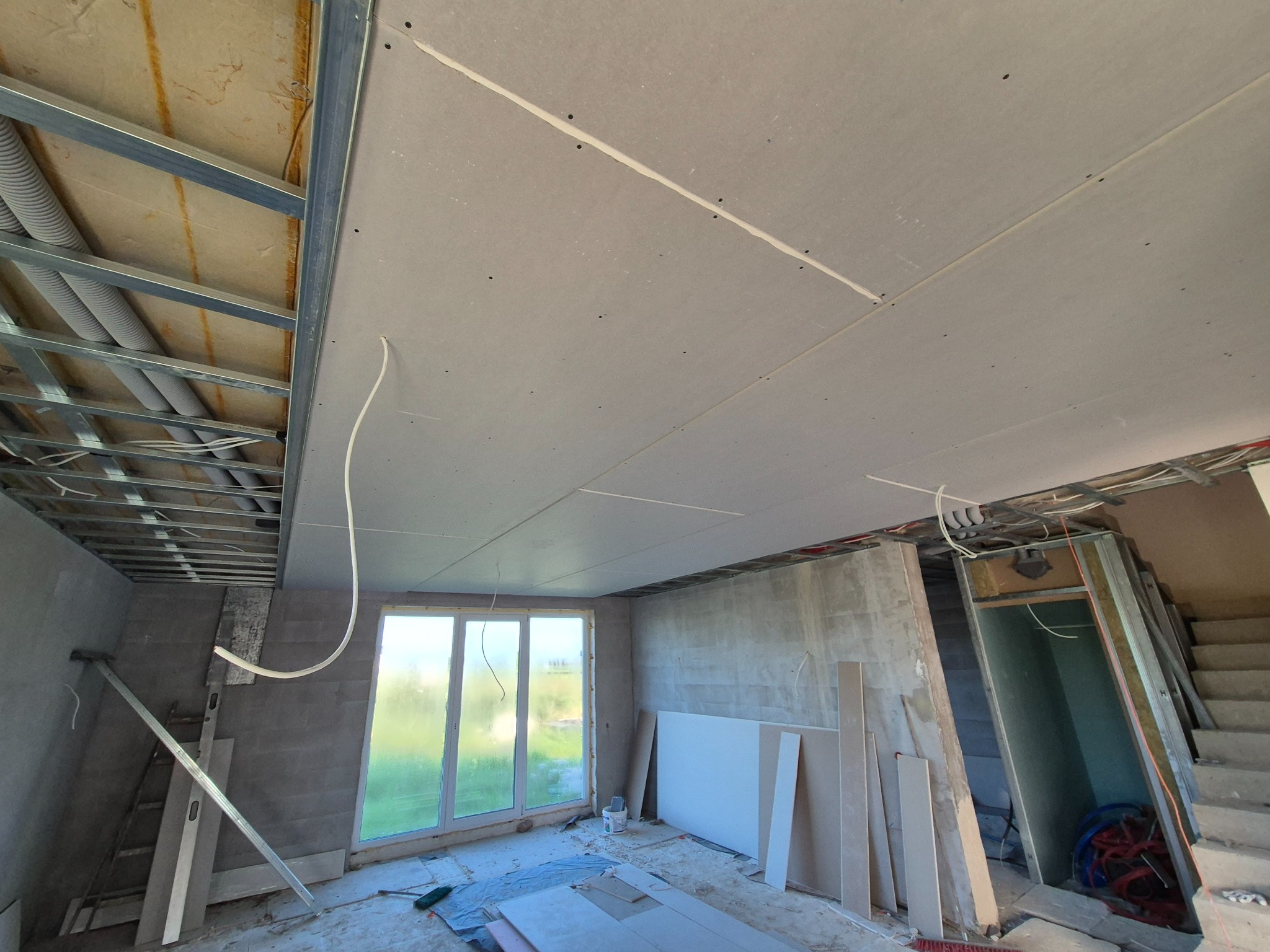 Montaż sufitu podwieszanego z płyt gipsowo-kartonowych na metalowej konstrukcji w surowym wnętrzu z widoczną instalacją elektryczną i wentylacyjną, okno z widokiem na zieleń w tle.