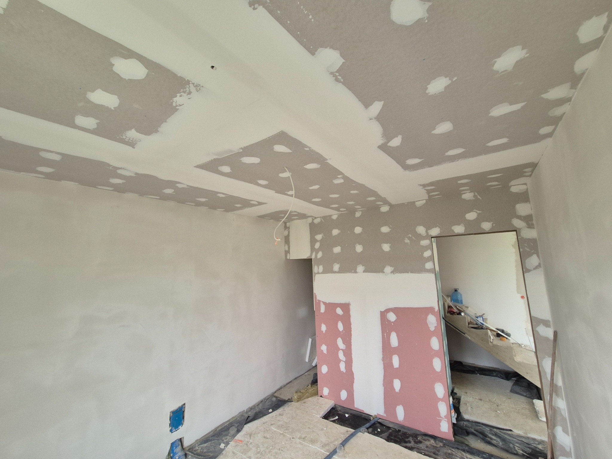 Wnętrze w trakcie prac wykończeniowych: sufit z płyt kartonowo-gipsowych z widocznymi łączeniami szpachlowanymi, ściany częściowo wygładzone, zabezpieczona folią podłoga i fragment różowej płyty...
