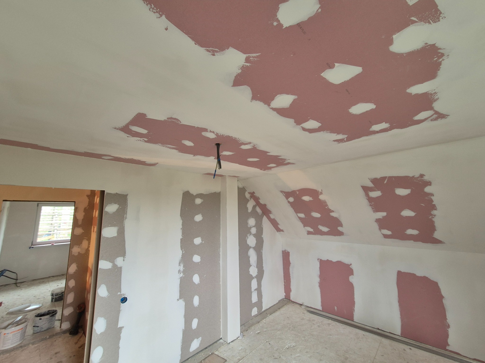 Poddasze w trakcie prac wykończeniowych: częściowo zagruntowane ściany i sufit z widocznymi fragmentami różowej płyty kartonowo-gipsowej, punktowe szpachlowanie w toku, widoczne kable oświetleniowe.