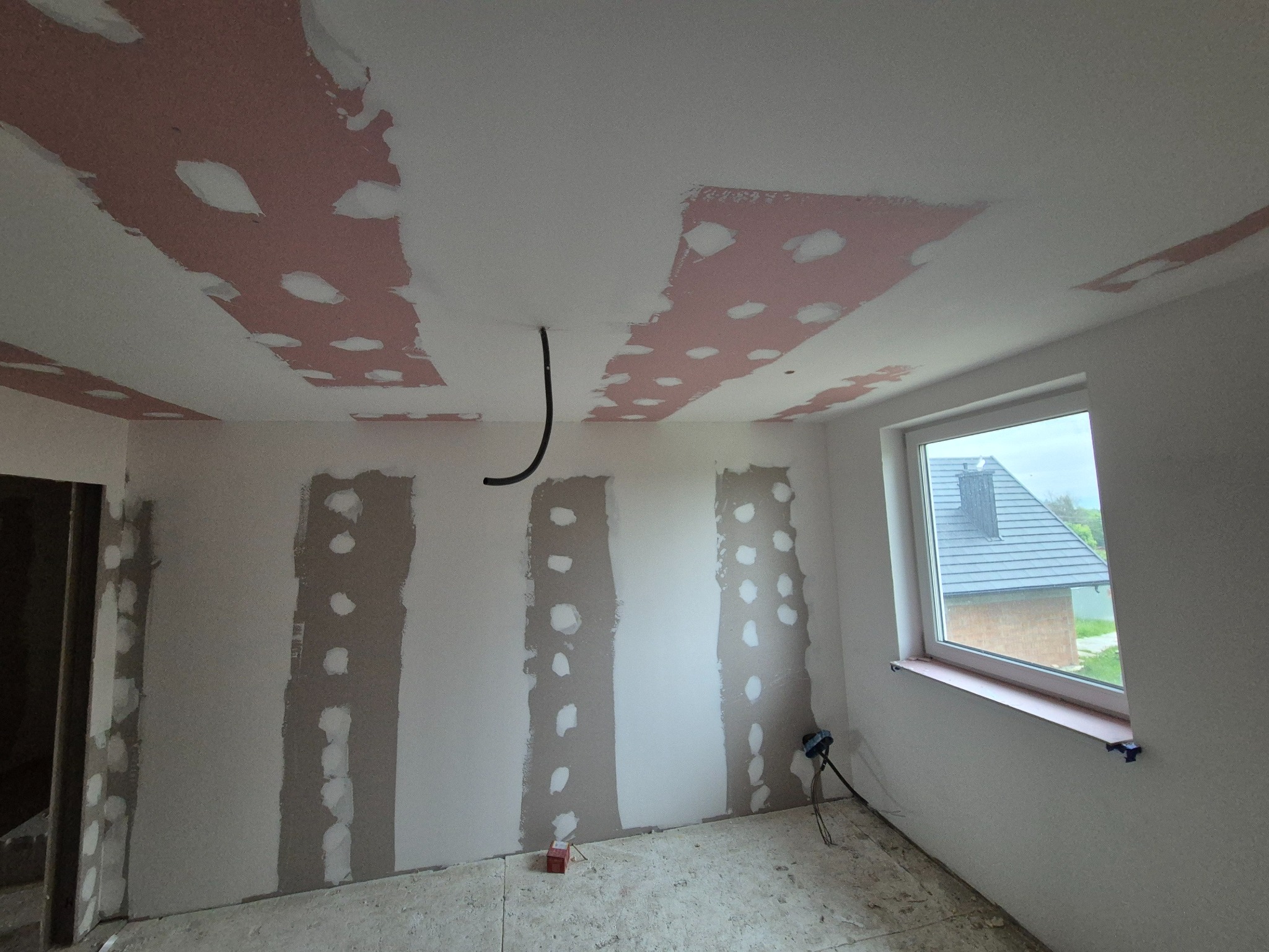 Wnętrze pokoju w trakcie remontu: częściowo położona płyta karton gips na suficie i ścianach, widoczne szpachlowane łączenia, kabel zwisający z sufitu, okno z widokiem na dom.