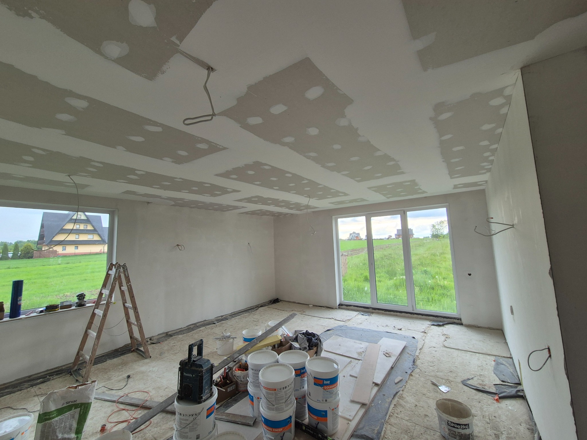 Pokój w trakcie prac wykończeniowych: sufit z widocznymi łączeniami płyt, ściany częściowo pokryte masą szpachlową, drabina przy oknie z widokiem na zielony krajobraz, wiadra z zaprawą i narzędzia...