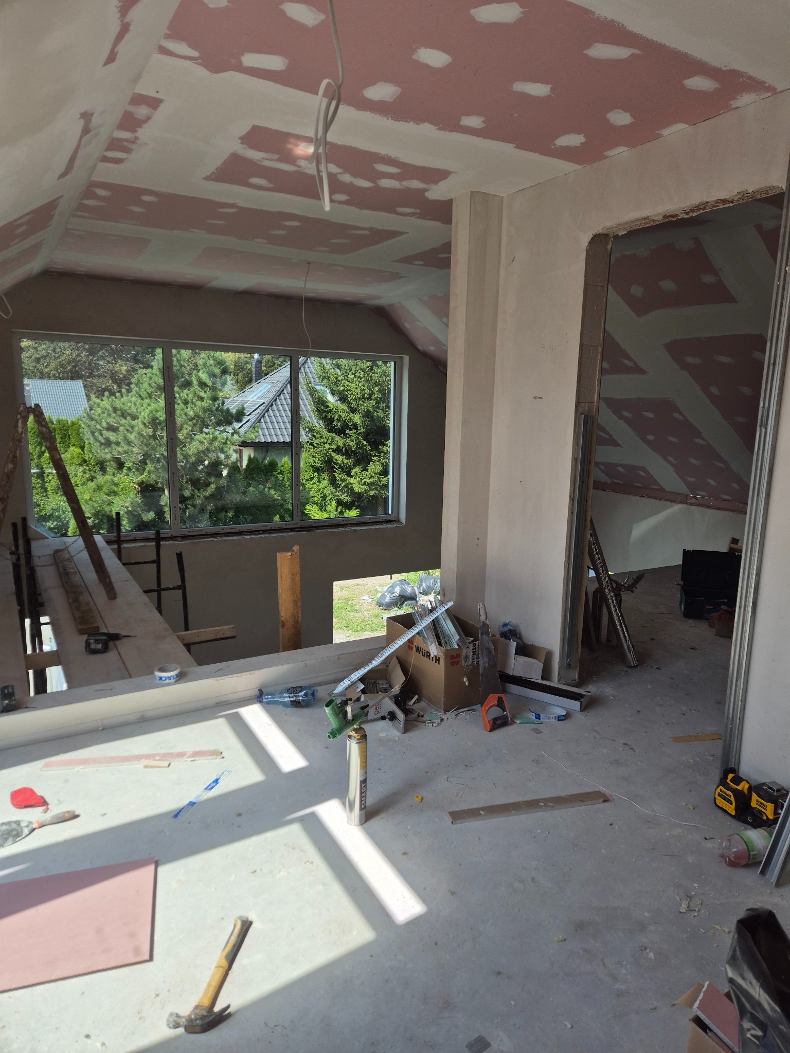 Poddasze w trakcie prac wykończeniowych: widoczne płyty na skosach sufitu z miejscami świeżo zaszpachlowanymi, okno z widokiem na zieleń, narzędzia i materiały budowlane porozrzucane na podłodze.